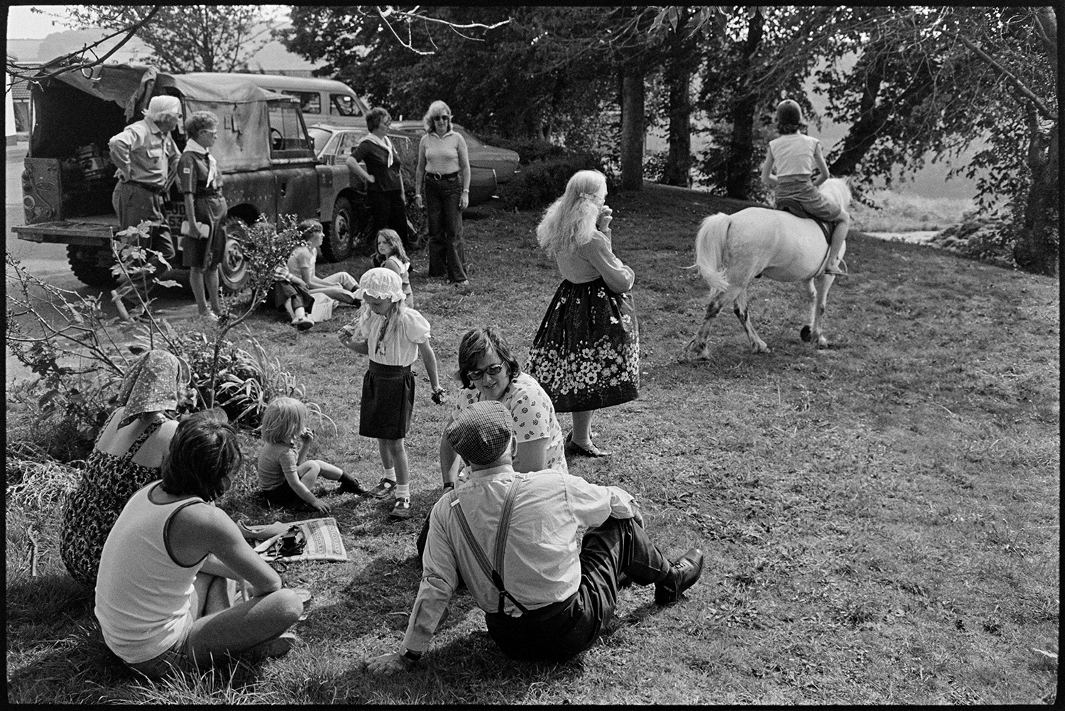 Barbeque fete in grounds of sheltered homes, people sitting out in the sun chatting. 
[Men, women and children sat on the grass at a fete in the grounds of Torridge View sheltered housing in Torrington. Men and women are talking and a child is having a pony ride.]