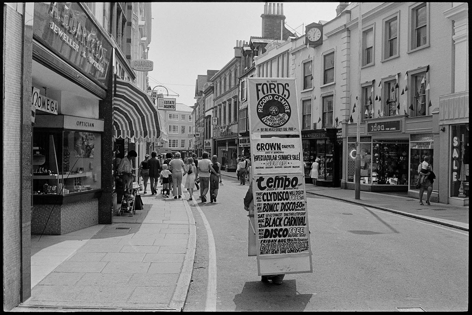 Street scene with sandwich man advertising record shop. 
[A person wearing a sandwich board advertising Fords Record Centre, Crown Carpets and various discos stood in Barnstaple High Street. Shoppers and various shop fronts can be seen including Idea for Living, Garnish and Winkles, Jewellers, A E Dark & Son , Jeweller and Optician and B C Estate Agents.]