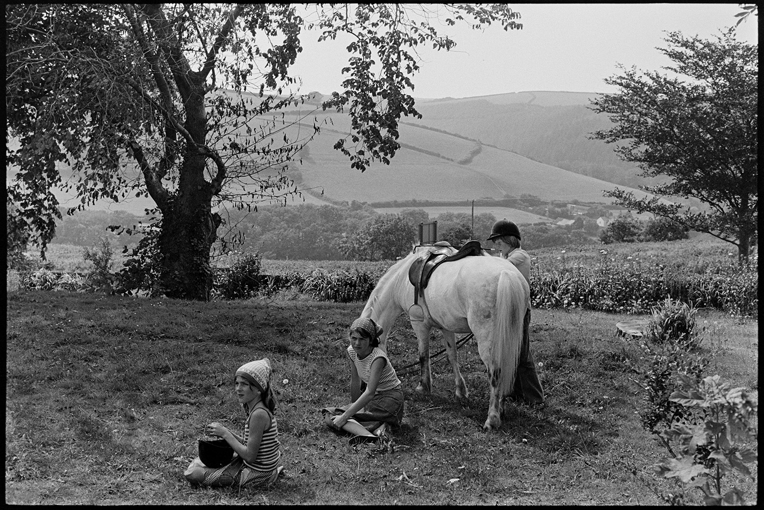 Scenes at opening of barbeque fete in grounds of sheltered homes, pony ride, roulette, etc. 
[Children at a fete at Torridge View sheltered housing in Torrington. They are sat on the grass with a pony, waiting to give pony rides in the grounds.]