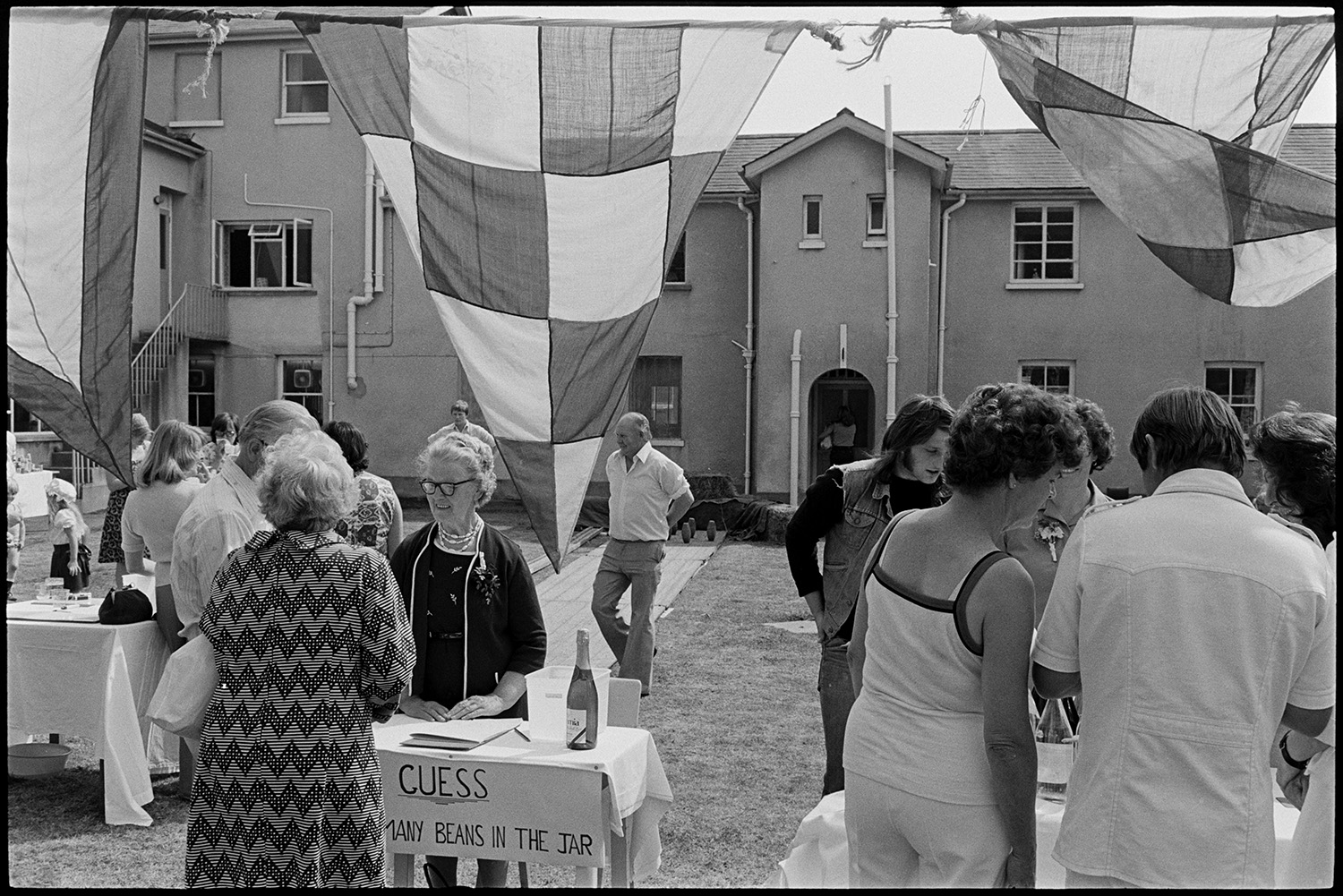 Scenes at opening of barbeque fete in grounds of sheltered homes, pony ride, roulette, etc. 
[Men and women looking at stall at a fete at Torridge View sheltered housing in Torrington. A woman is taking part in a game to guess how many beans are in a jar. A skittle alley can also be seen in the background.]