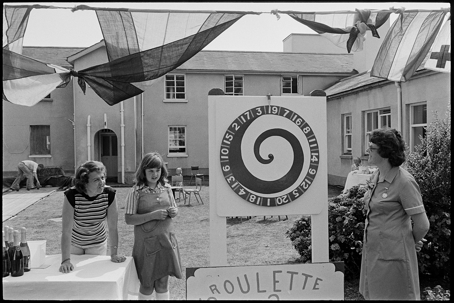 Scenes at opening of barbeque fete in grounds of sheltered homes, pony ride, roulette, etc. 
[A woman running a roulette game at a fete at Torridge View sheltered housing in Torrington. A girl and another woman are watching. In the background a man is setting up a skittles game.]