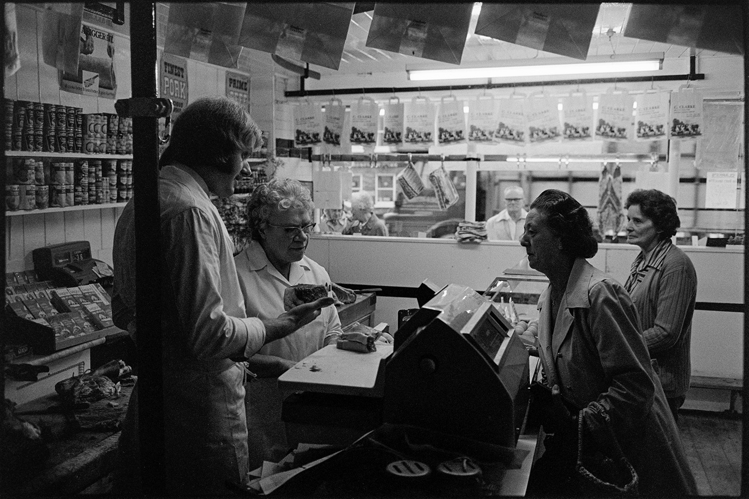 Interior of butcher's shop with customers. 
[A woman buying cuts of meat in a butcher's shop in South Molton. A man and woman behind the counter are weighing the cuts of meat on the shop till. Various tins are displayed on shelves behind the counter.]