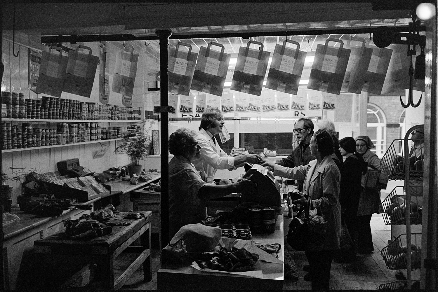 Interior of butcher's shop with customers. 
[A man and woman serving customers in a butcher's shop in South Molton. Behind the till various tins of food are on display and in the foreground baskets with fresh fruit and vegetables are visible.]