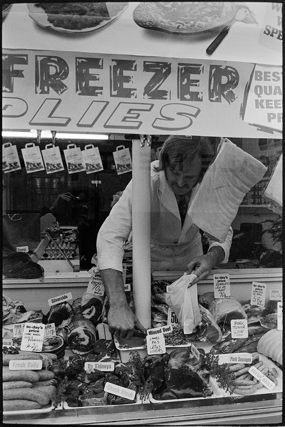 Interior of butcher's shop with customers. 
[A butcher bagging up cuts of meat from a window display in his butcher's shop for a customer, in South Molton.]