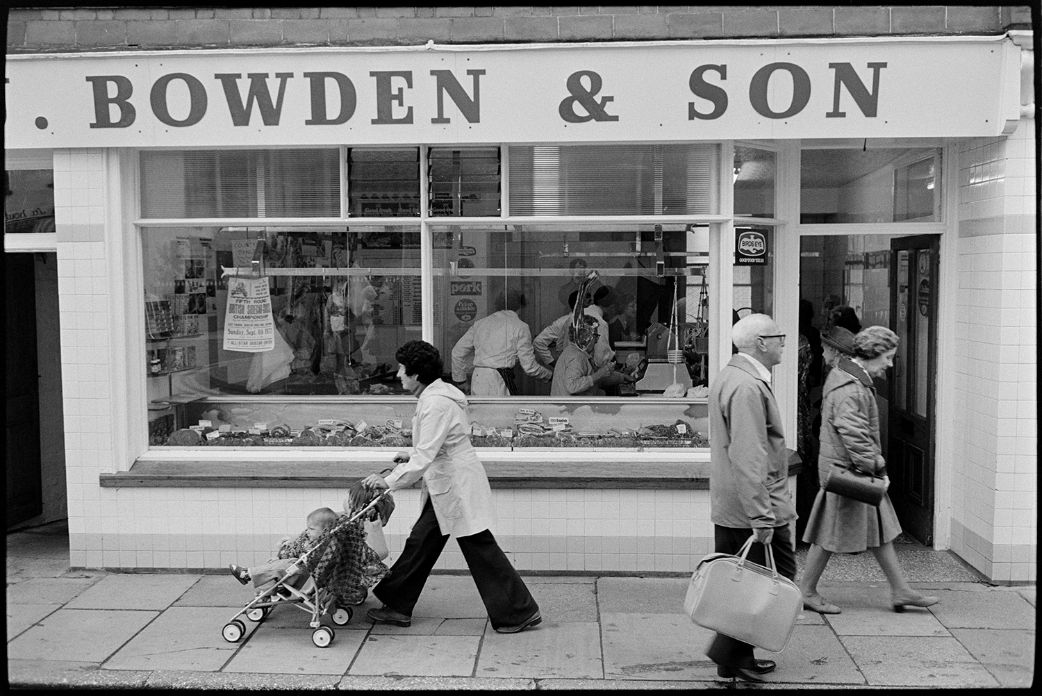 Interior of butcher's shop with customers, also front of shop. 
[The shop front window of Bowden & Son butcher's shop in South Molton. People are walking past, including a woman pushing a pram. Various cuts of meat are displayed and hung up in the window and staff can be seen serving customers.]