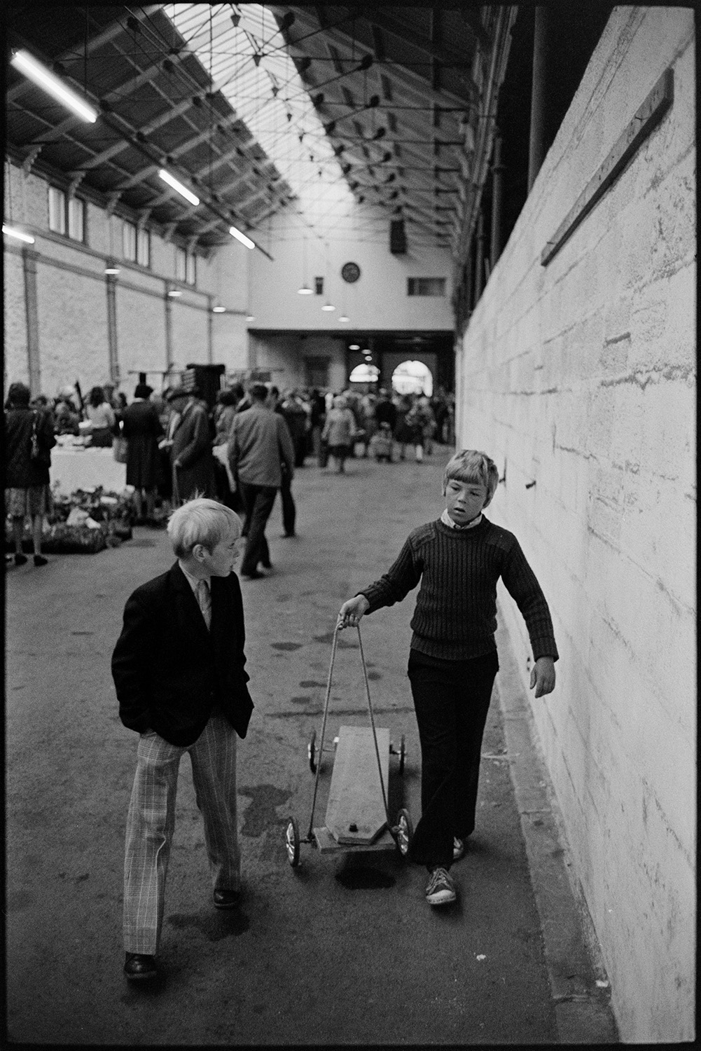 Market hall with stalls, fruit, vegetables and flowers, customers. 
[Two boys in South Molton Pannier Market with a soapbox or scooter. Men and women can be seen shopping at stalls in the market in the background.]