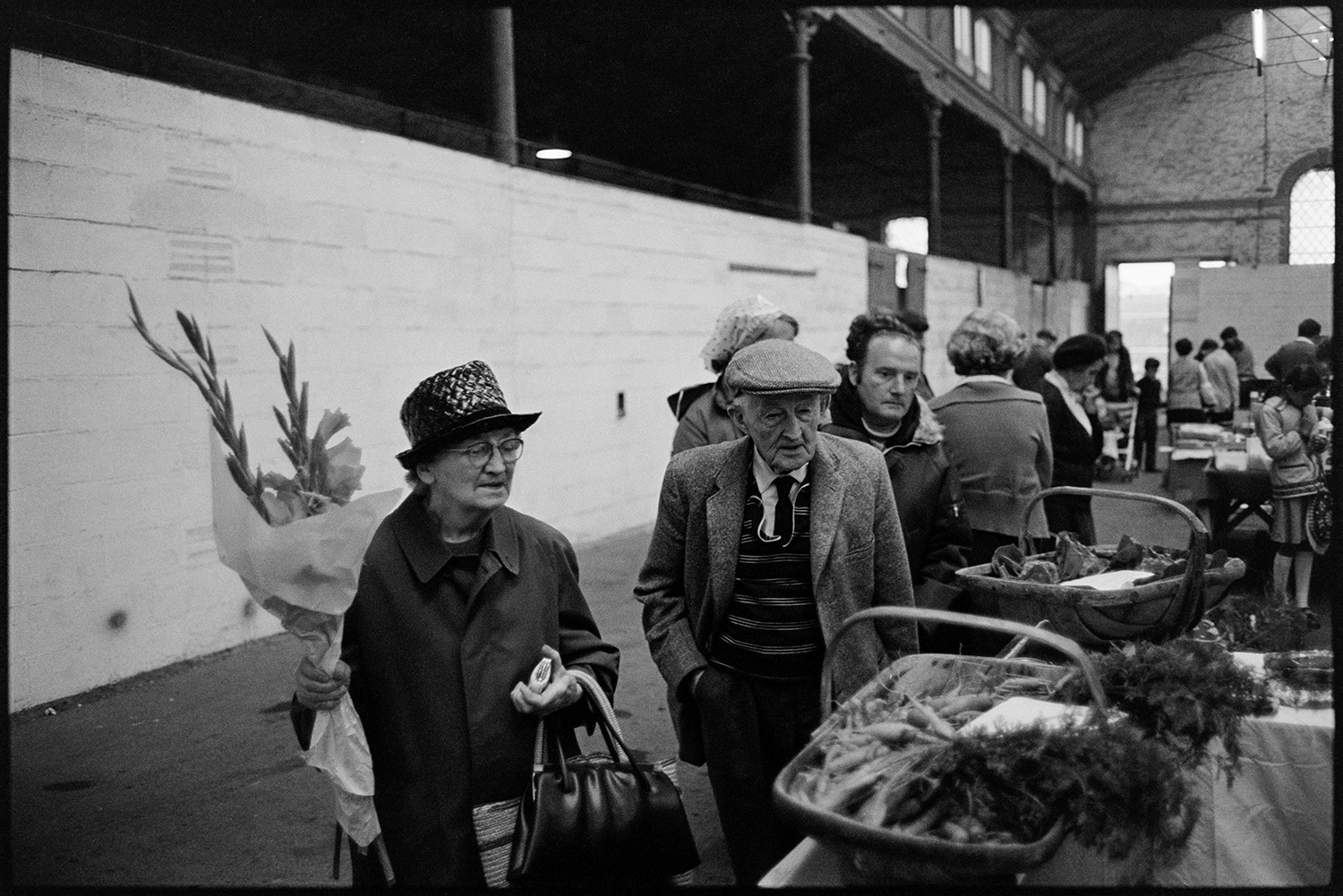 Market hall with stalls, fruit, vegetables and flowers, customers. 
[A man and woman looking at vegetables, including carrots, on a market stall at South Molton Pannier Market. The woman is holding a bunch of flowers and bags. Other customers can be seen in the background.]