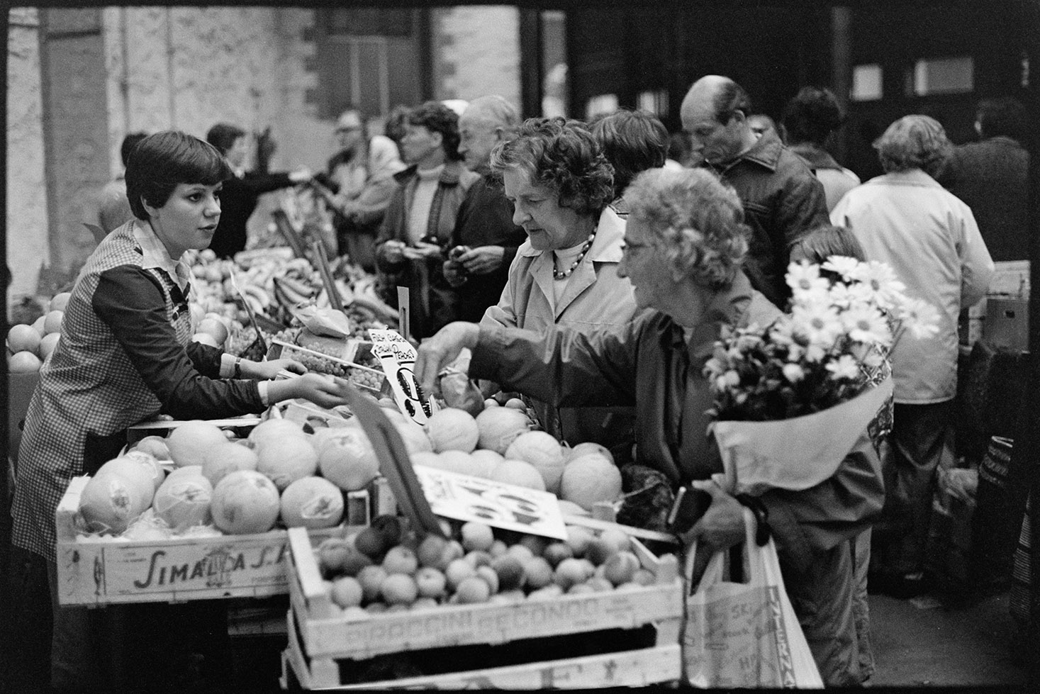 Market hall with stalls, fruit, vegetables and flowers, customers. 
[Two women looking at a fruit stall and talking to the market trader at South Molton Pannier Market. Melons are visible on the stall and other customers can be seen in the background. One of the women is holding a bunch of flowers.]