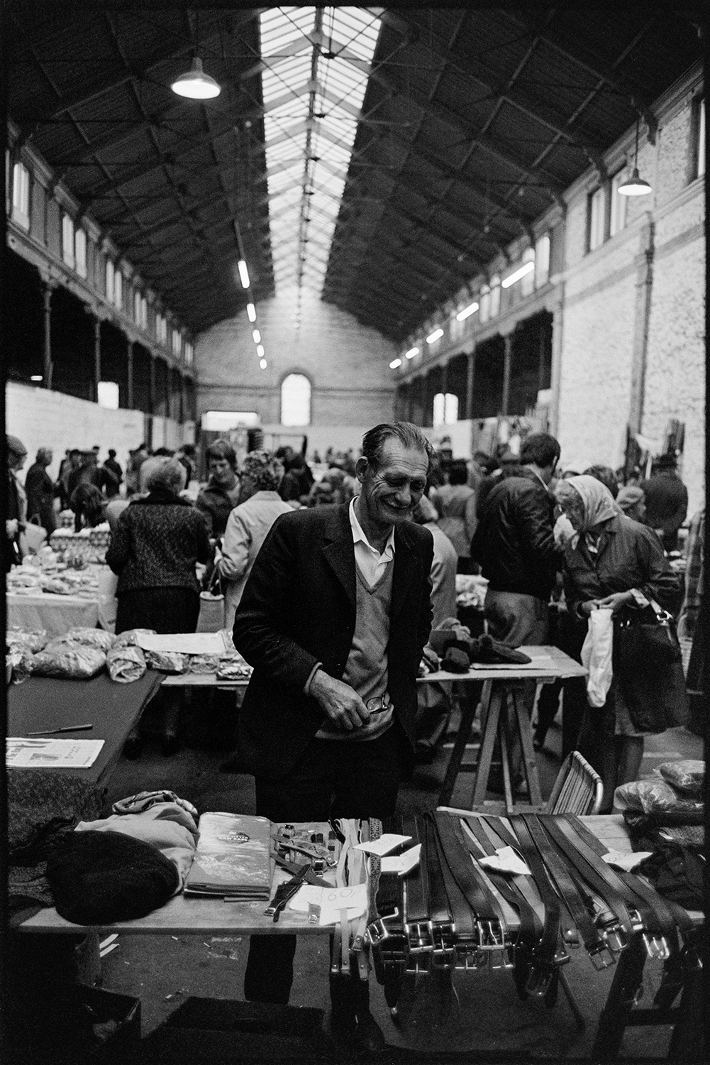 Market hall with stalls, fruit, vegetables and flowers, customers. 
[A man at his market stall selling leather goods at South Molton Pannier Market. Belts and watch straps are visible on the stall.]