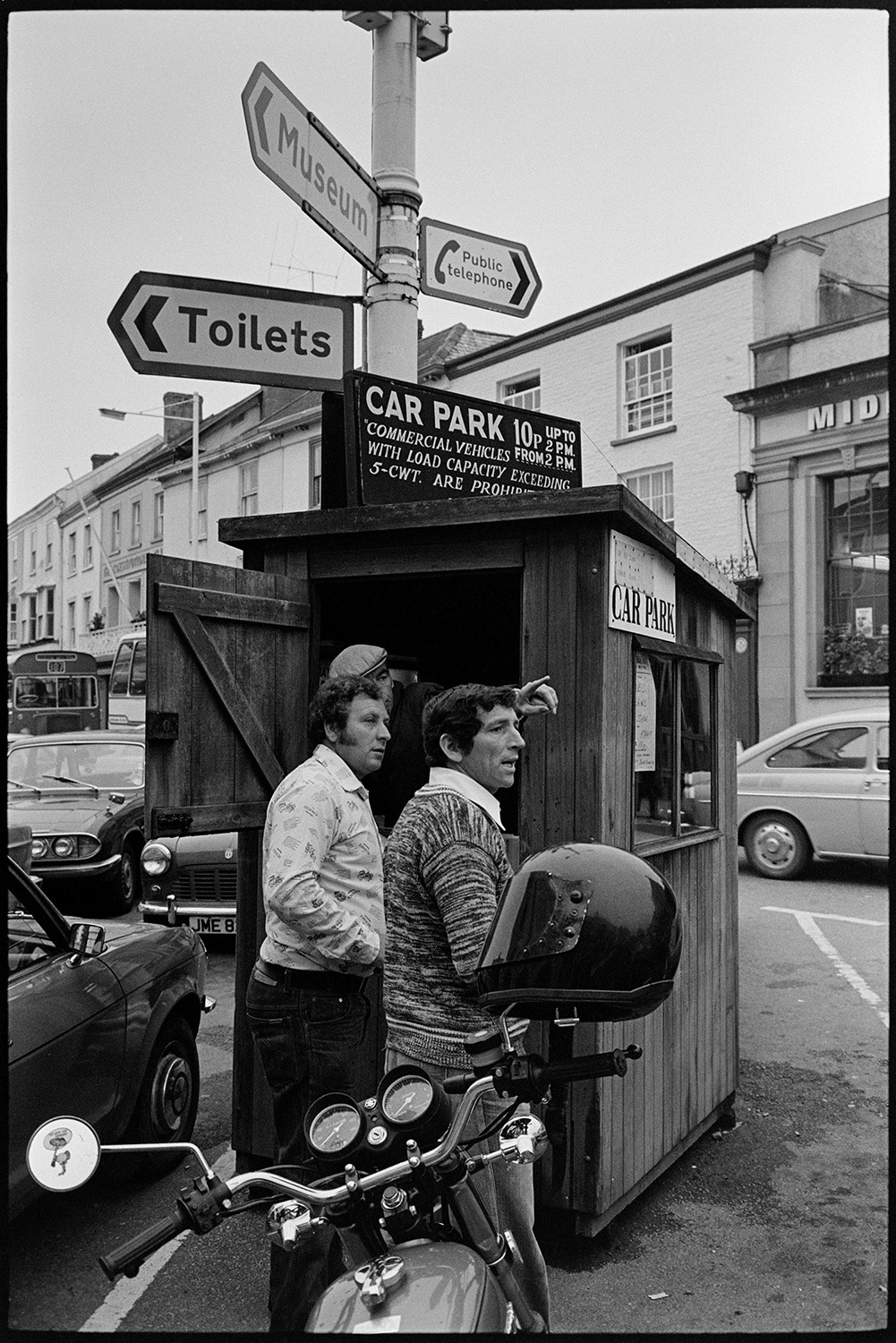 Car park attendant and hut in town square with road signs. 
[A car park attendant giving directions to two men  from his wooden hut in the car park in South Molton town square. A signpost is above the wooden hut.]