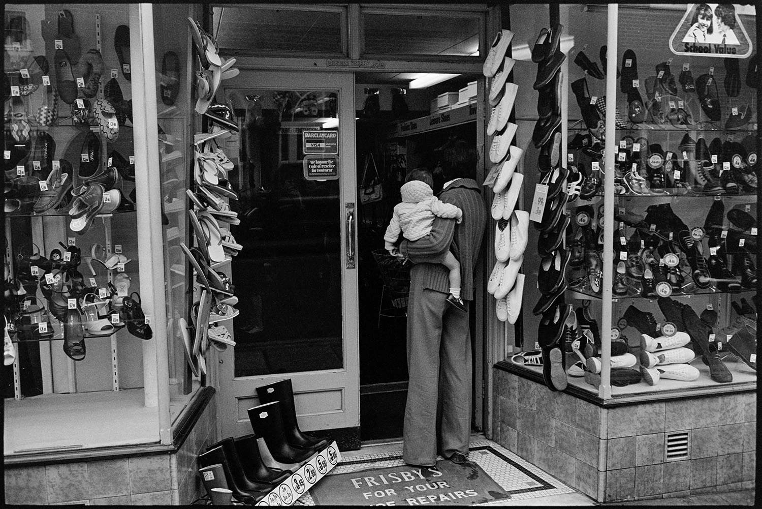 Front of shoe shop with woman and child. 
[A woman and child looking in the front door of Frisbys shoe shop in South Molton. A variety of shoes and wellington boots are displayed in the shop window.]