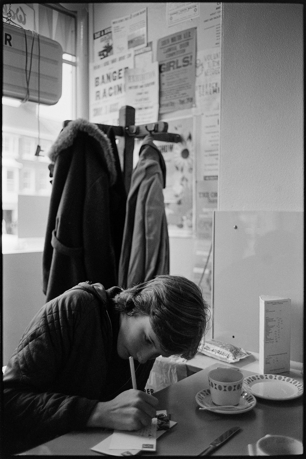A teenager in a café writing a post card. A coat stand and posters are visible in the background.