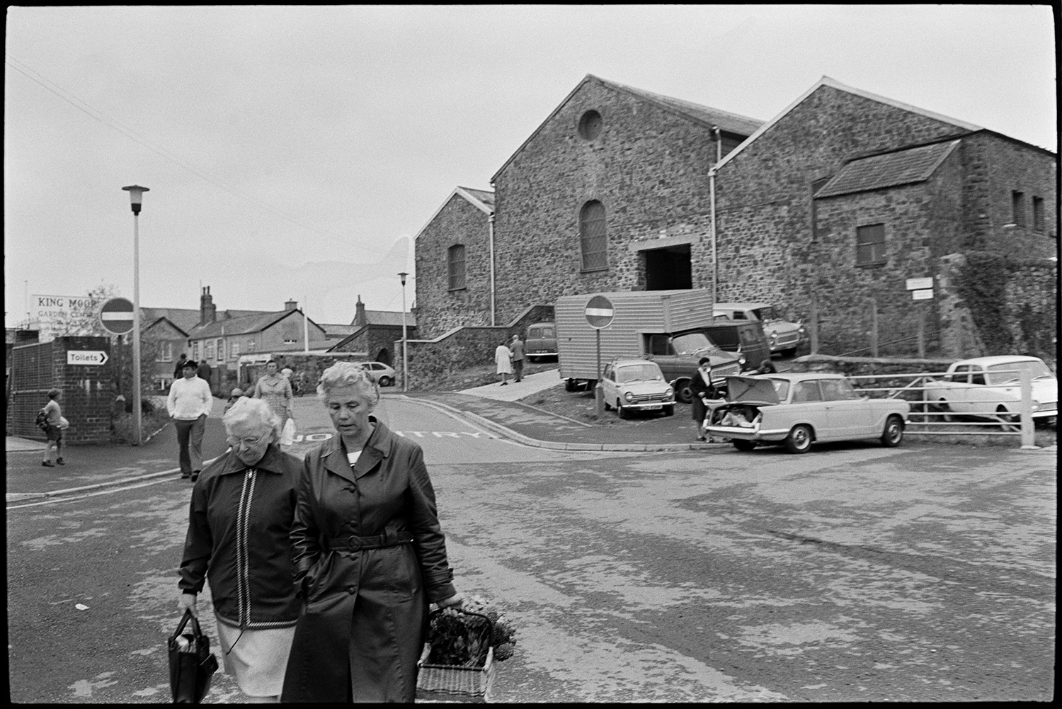 Market scenes. 
[Two women walking away from the back of the South Molton Pannier Market building. They are holding bags with produce they have bought. Various parked cars and a lorry can be seen by the market entrance.]