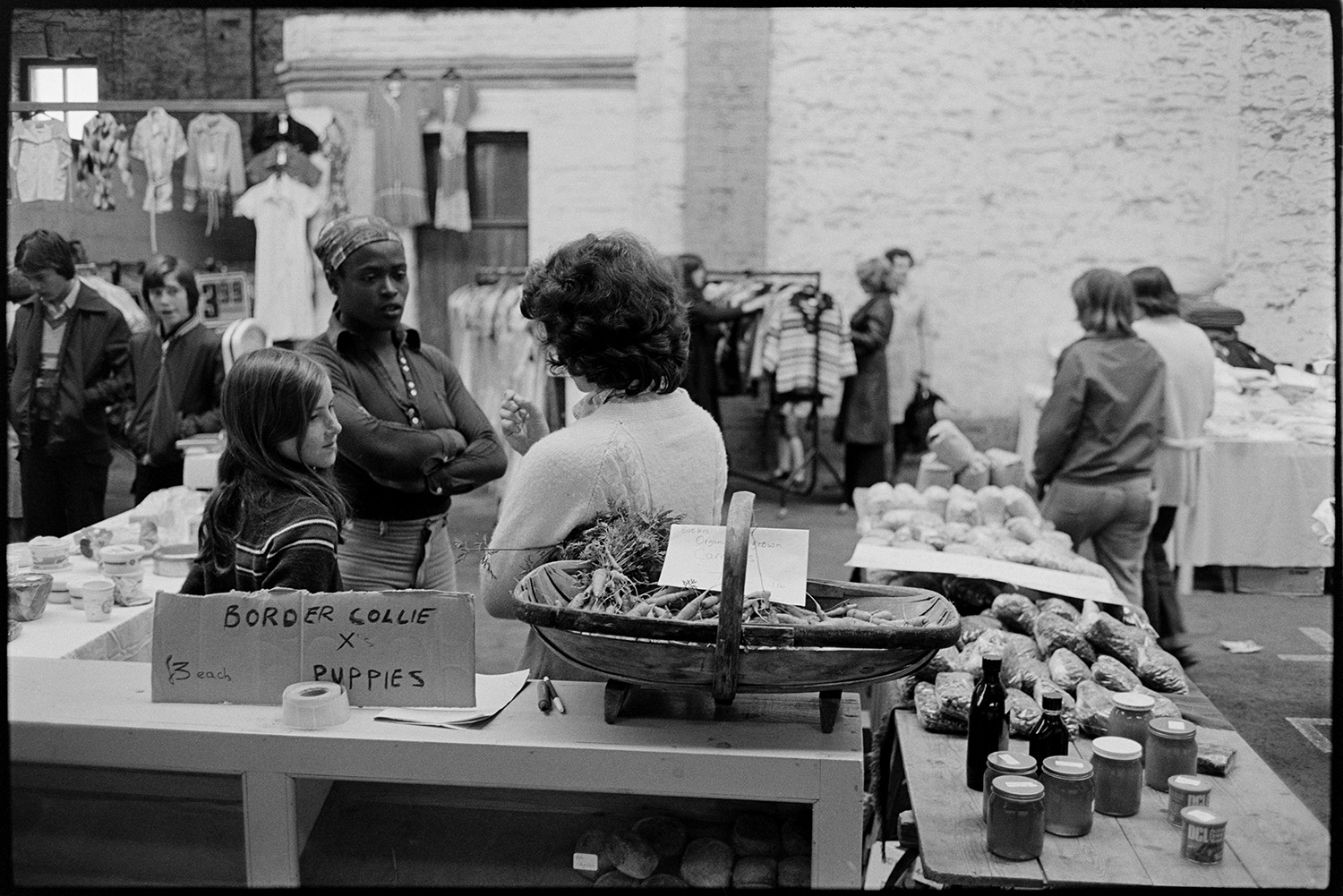 Market scenes. 
[Two women and a girl talking behind a market stall at South Molton Market. Jars, packets of food and a trug with carrots can be seen on the stall. There is also a sign advertising Border Collie cross puppies. Customers and other market stalls, including a clothes stall, can be seen in the background.]