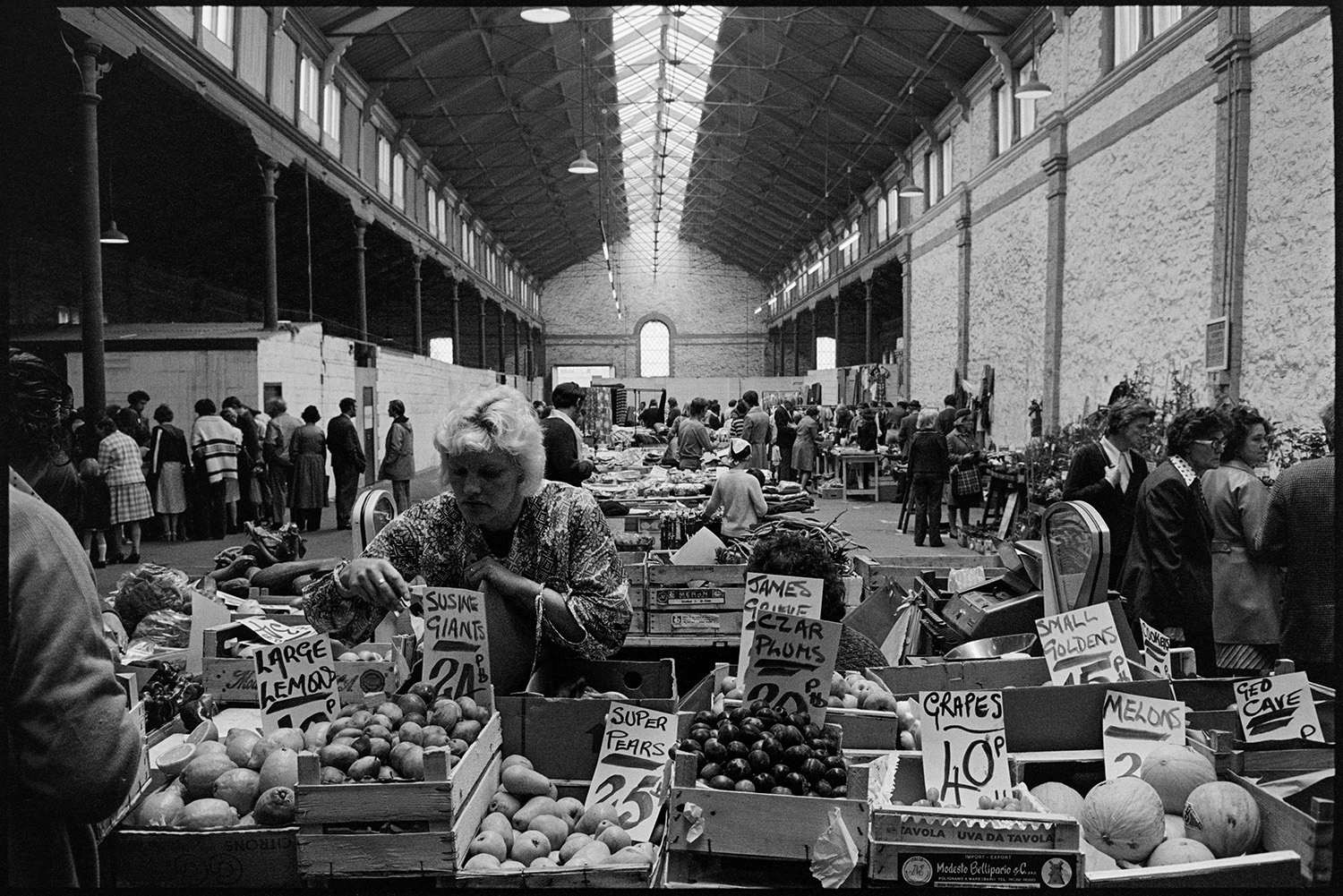 Cloth stall in market. 
[A woman running a fruit stall at South Molton Pannier Market. The stall has lemons, pears, plums and melons, amongst other fruit. Shoppers can be seen looking at other stalls in the background.]