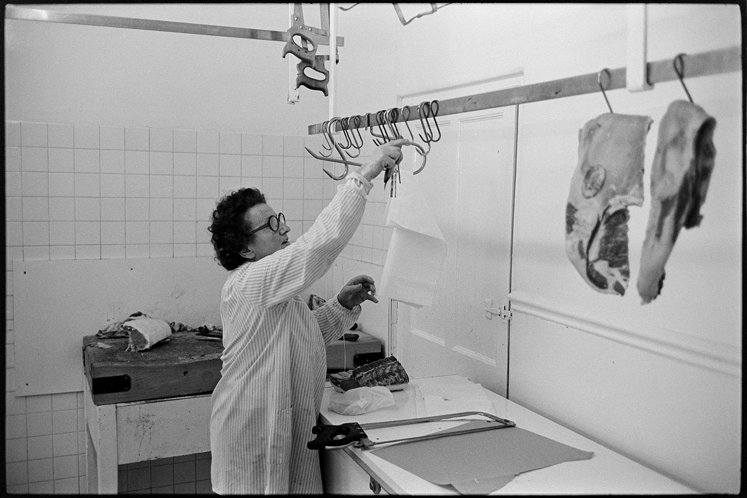 Butchers shop, serving customer. 
[A woman hanging up meat hooks in a butcher's shop in South Molton. Some of the hooks have cuts of meat hanging from them.]