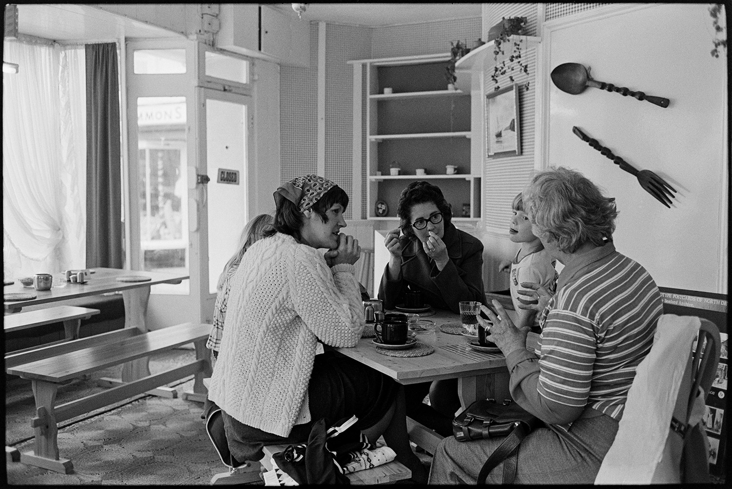 Customers in restaurant café. 
[A group of women and children having drinks in a café in South Molton. A bench table is behind them and a decorative spoon and fork are hanging on the wall.]