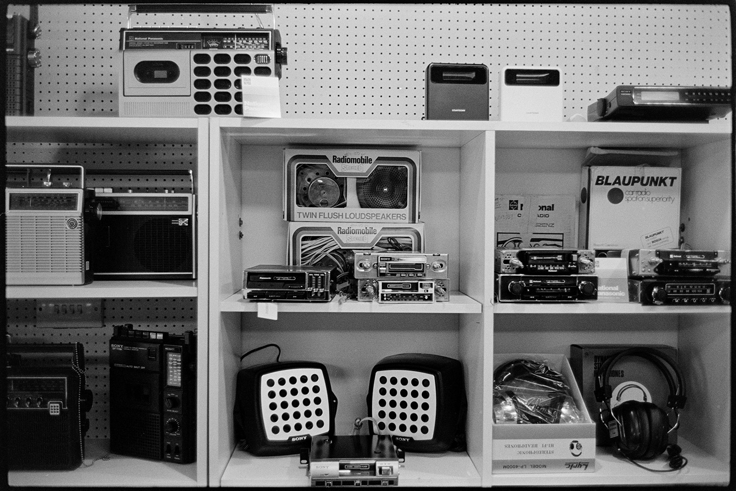 Goods on shelf in Radio, Hi Fi and television shop. 
[The interior of J P Williams electrical shop in South Molton. Radios, tape recorders, speakers and headphones are on display amongst other items,]