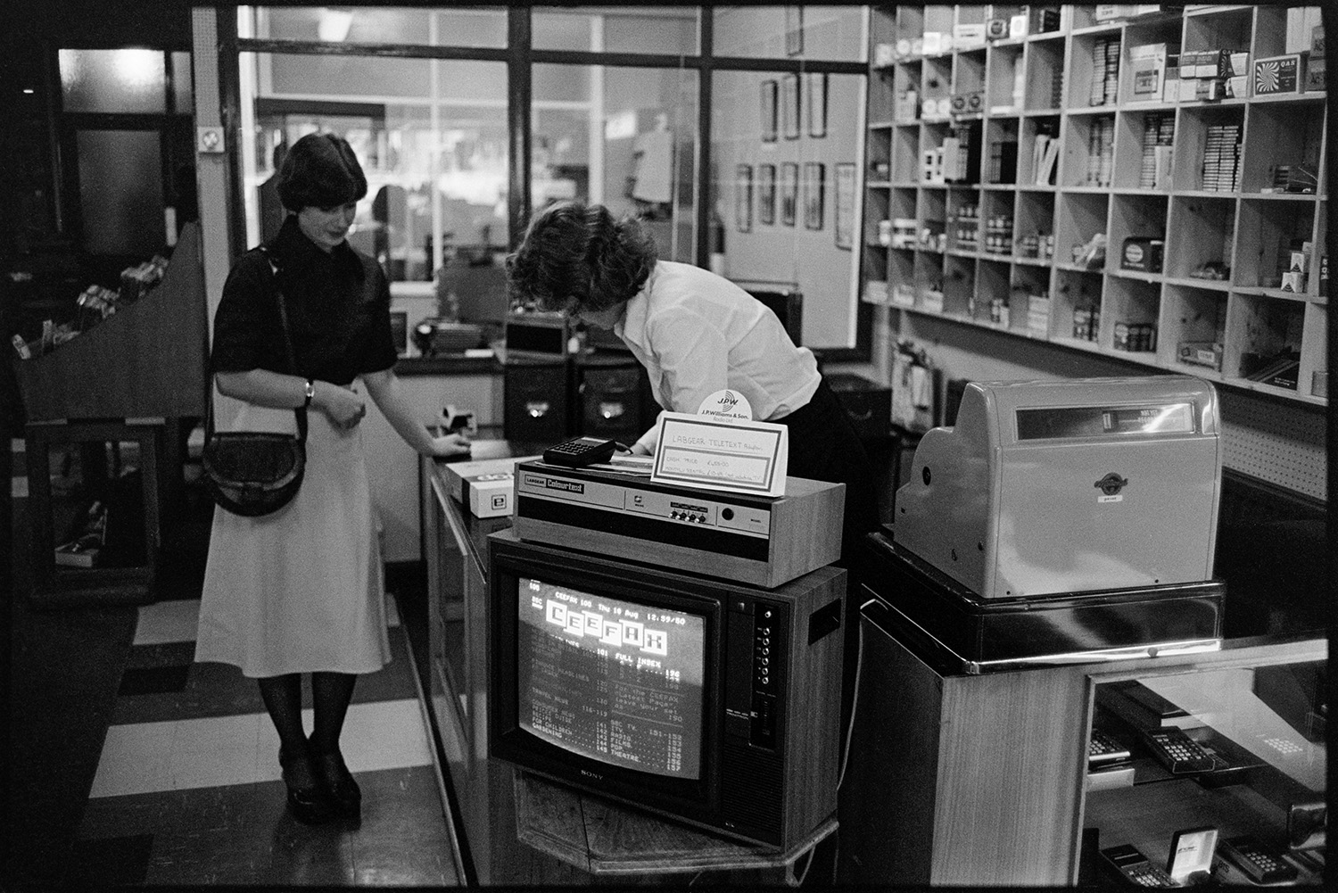 Goods on shelf in Radio, Hi Fi and television shop. 
[A woman serving a customer at the counter of J P Williams electrical shop in South Molton. A television is displayed by the counter with a Ceefax page on show. Various other goods are stacked on shelves behind the counter.]