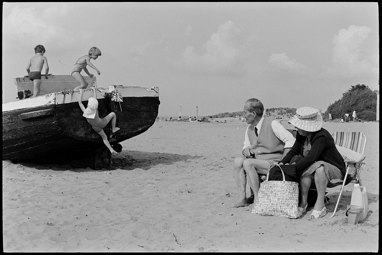 Beach with children playing on beached barge. 
[A man and woman sat on garden chairs on Instow beach. They are watching three children playing on a beached barge on the sand.]
