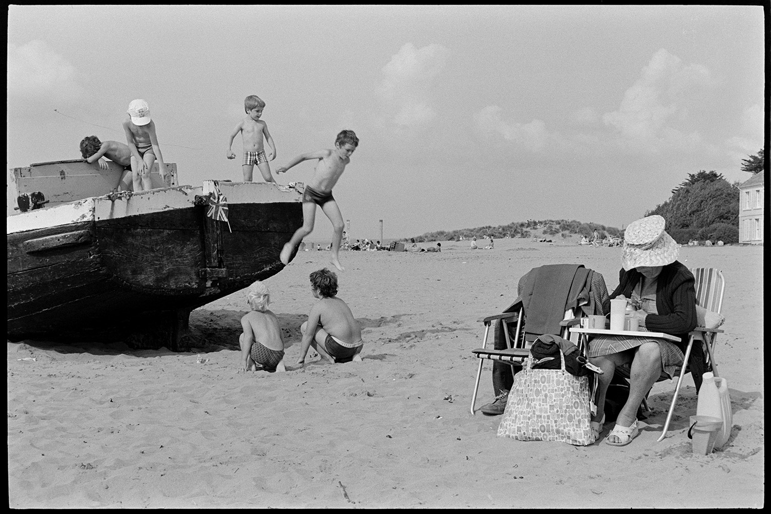 Beach with children playing on beached barge. 
[A woman sat on a garden chair at Instow beach. She is making drinks on a tray on her lap. Six children are playing on a beached barge on the sand nearby.]