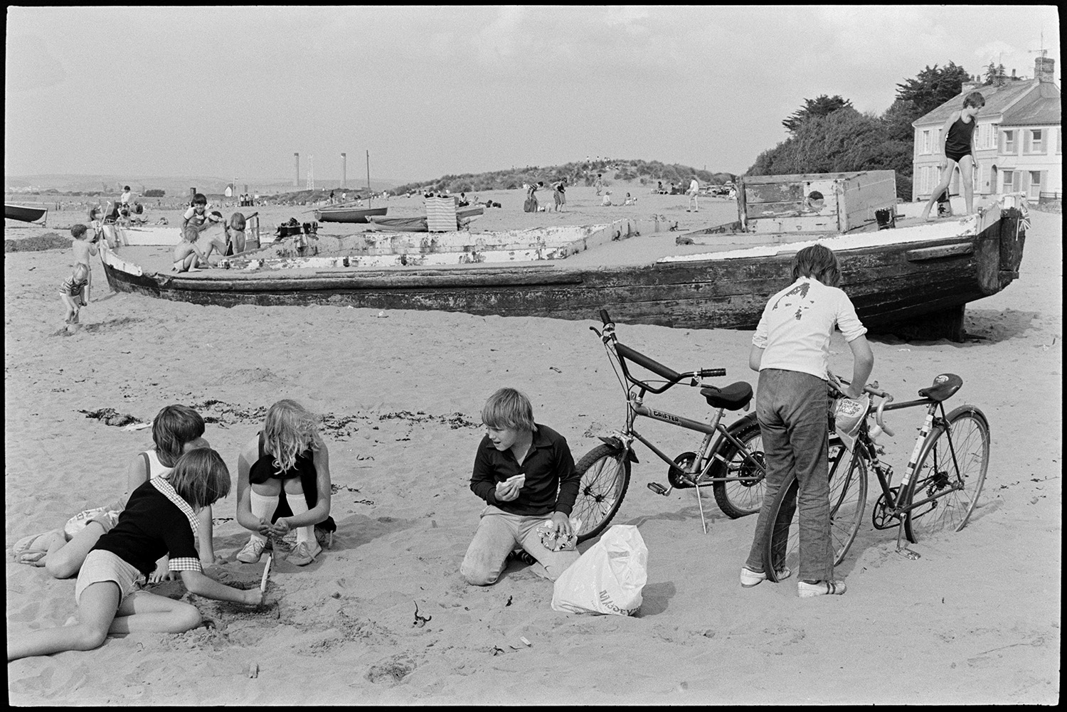 Beach with children playing on beached barge. 
[Children playing on the beach at Instow. One boy is eating a sandwich and another is looking in a bag attached to a bicycle. In the background children are playing on a beached barge. ]