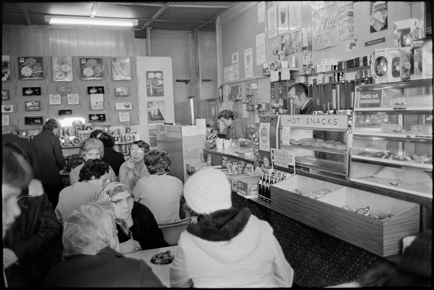 Cafe with bar and customers. 
[People sat in a cafe in Okehampton. A man and woman are preparing food and drink behind the counter. Bottled drinks and hot snacks are displayed on the counter and in the background posters or prints are hung on the wall.]