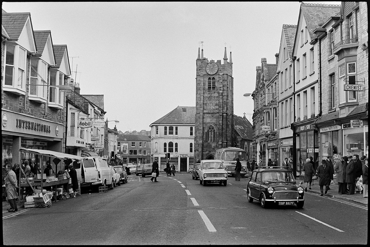 Town centre with cars, taken to compare with old photos. 
[The town centre of Okehampton with church tower and shops. Shop fronts include the International Stores, Chemist and Gordon Vick & Partners, estate agents. People can also be seen shopping at a market stall on the side of the street.]
