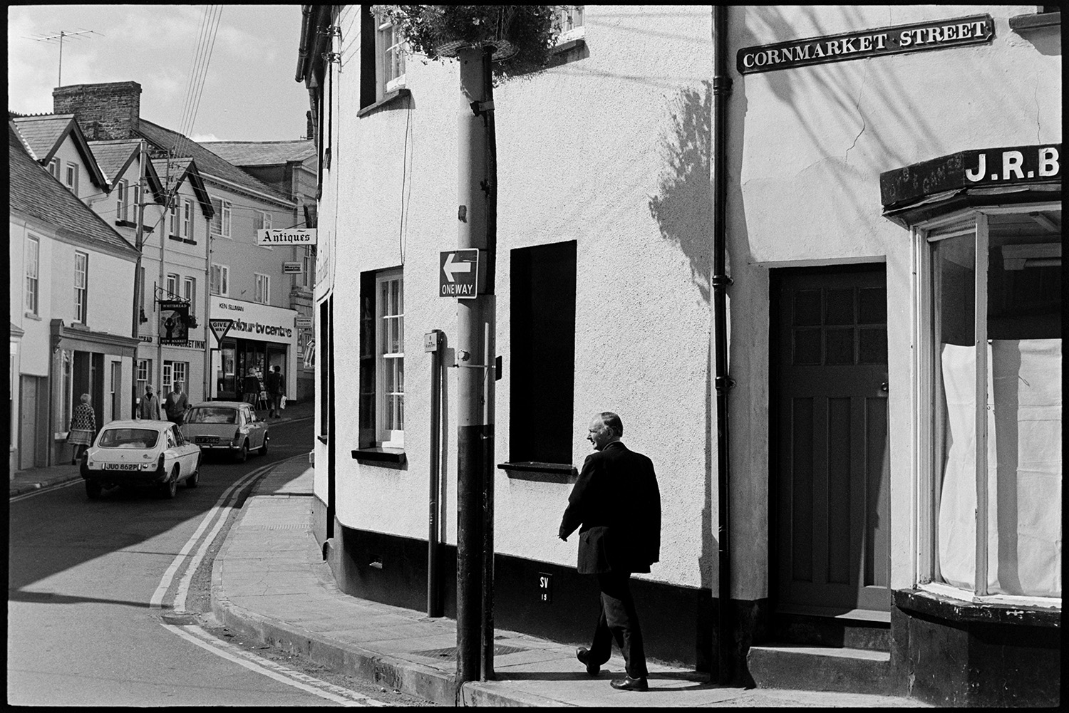 Street scenes with postwoman road sweeper and passers by. 
[A man walking along Cornmarket Street in Torrington. Cars, shoppers and shop front can be seen further up the street, including an Antiques shop Newmarket Inn and a TV Centre.]