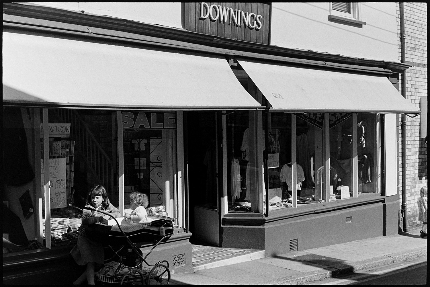Front of clothes shop and haberdashers. 
[The shop front of Downings clothes and haberdashery shop in Torrington. A child is stood outside with a pram and baby. Clothes are display in the shop window.]