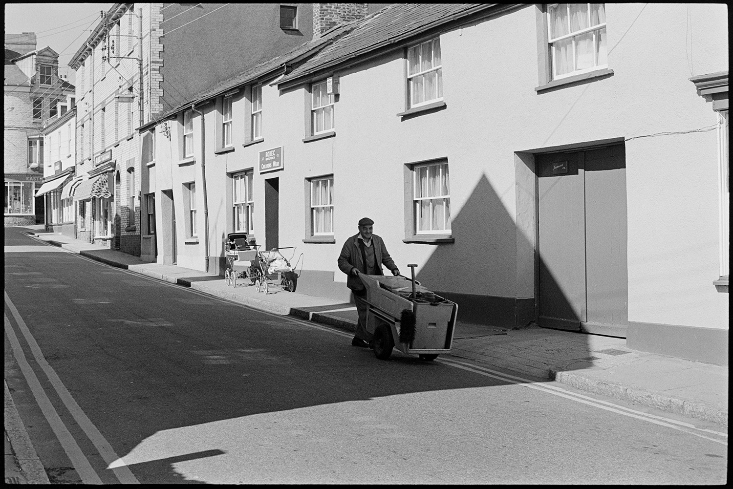 Street scenes with postwoman road sweeper and passers by. 
[A man pushing a road sweeper or street cleaner along a street in Torrington. Shop awnings can be seen in the background and two prams are parked outside Robec children's wear shop.]