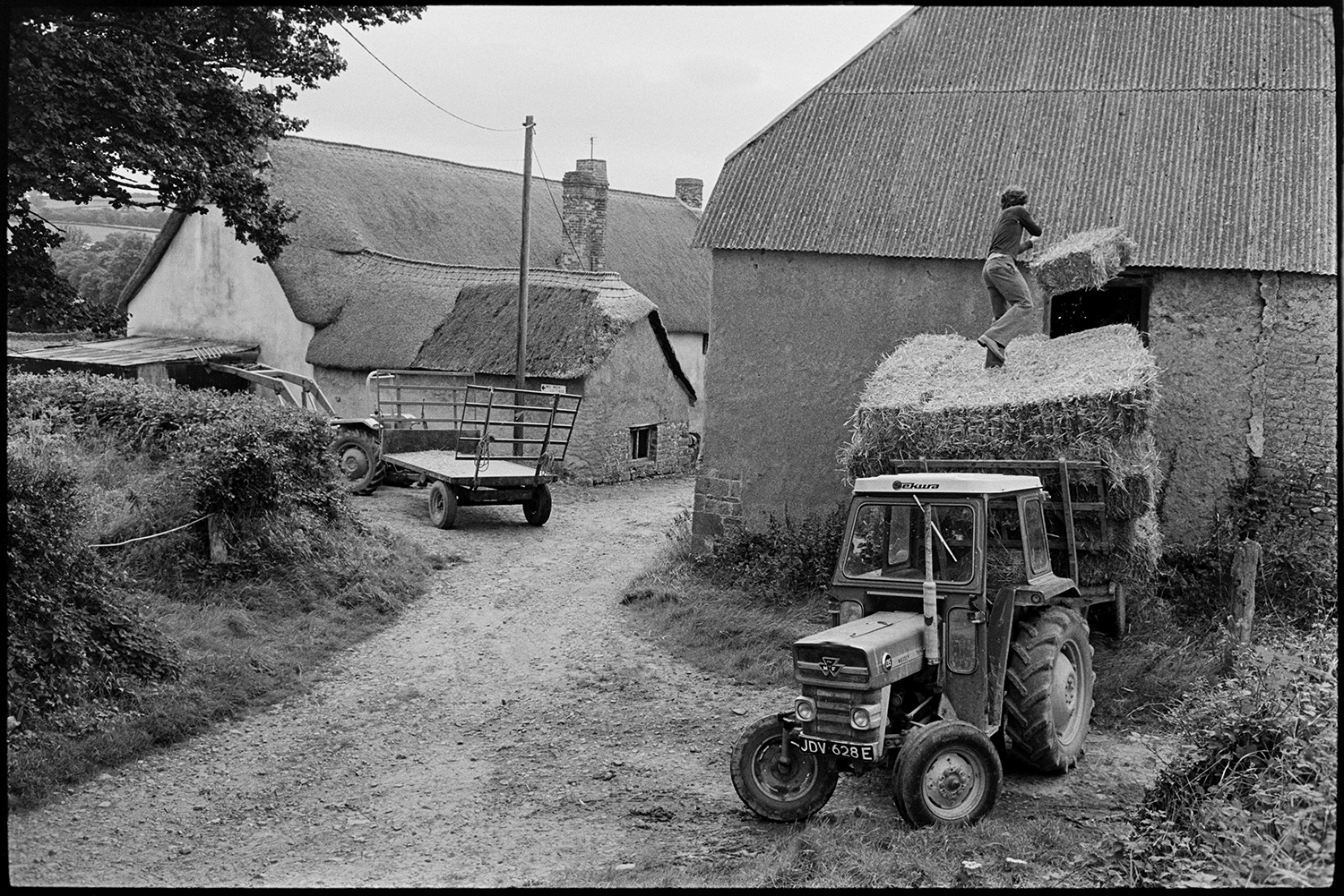Loading hay into barn tallet from tractor and trailer. 
[A man loading hay bales into a barn tallet from a tractor and trailer at Higher Week, Iddesleigh. Another tractor and trailer and thatched farmhouse are visible in the background.]