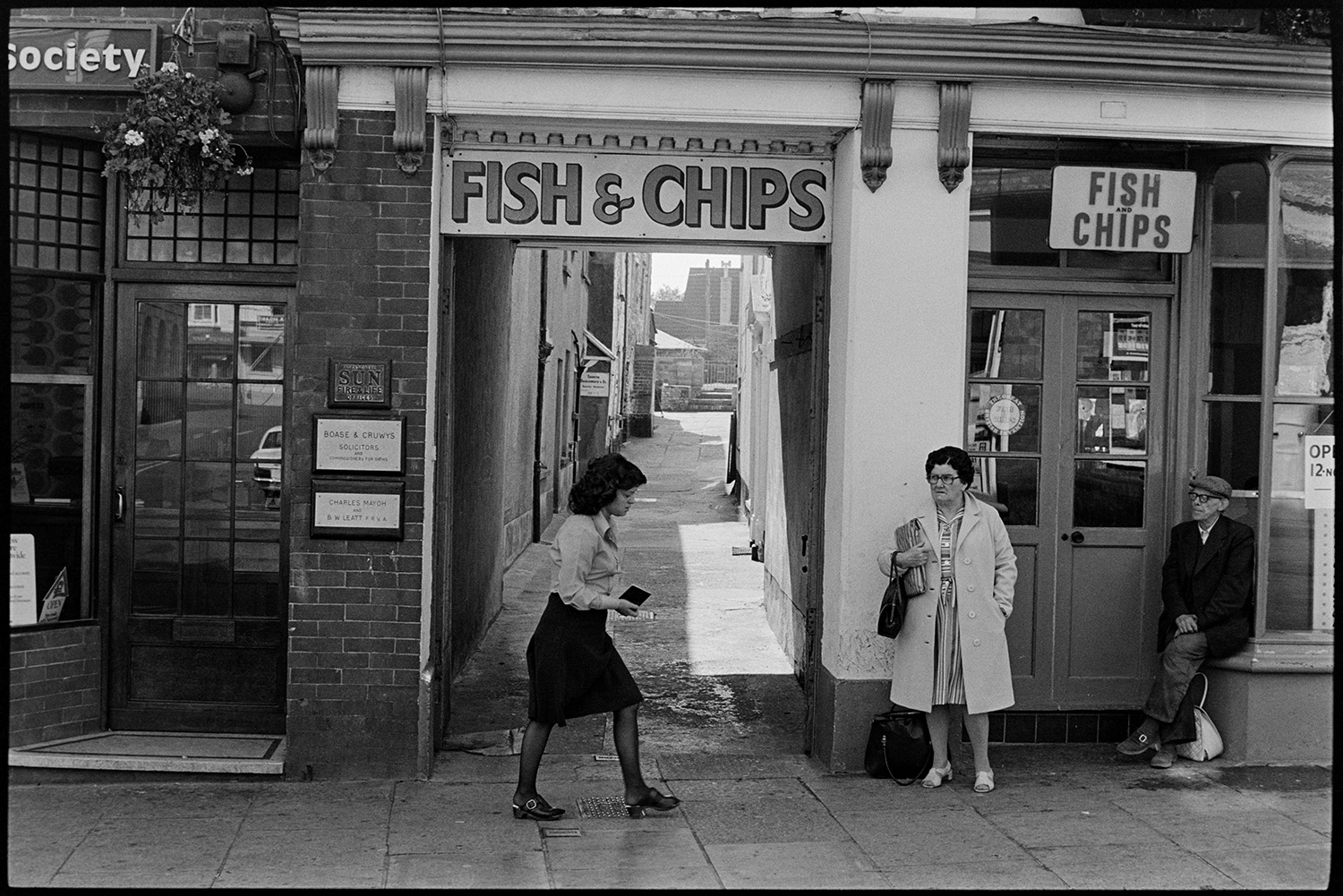 Front of fish and chip shop. 
[A man and woman waiting outside a fish and chip shop in Torrington. Another woman is walking past the alley next to the shop.]