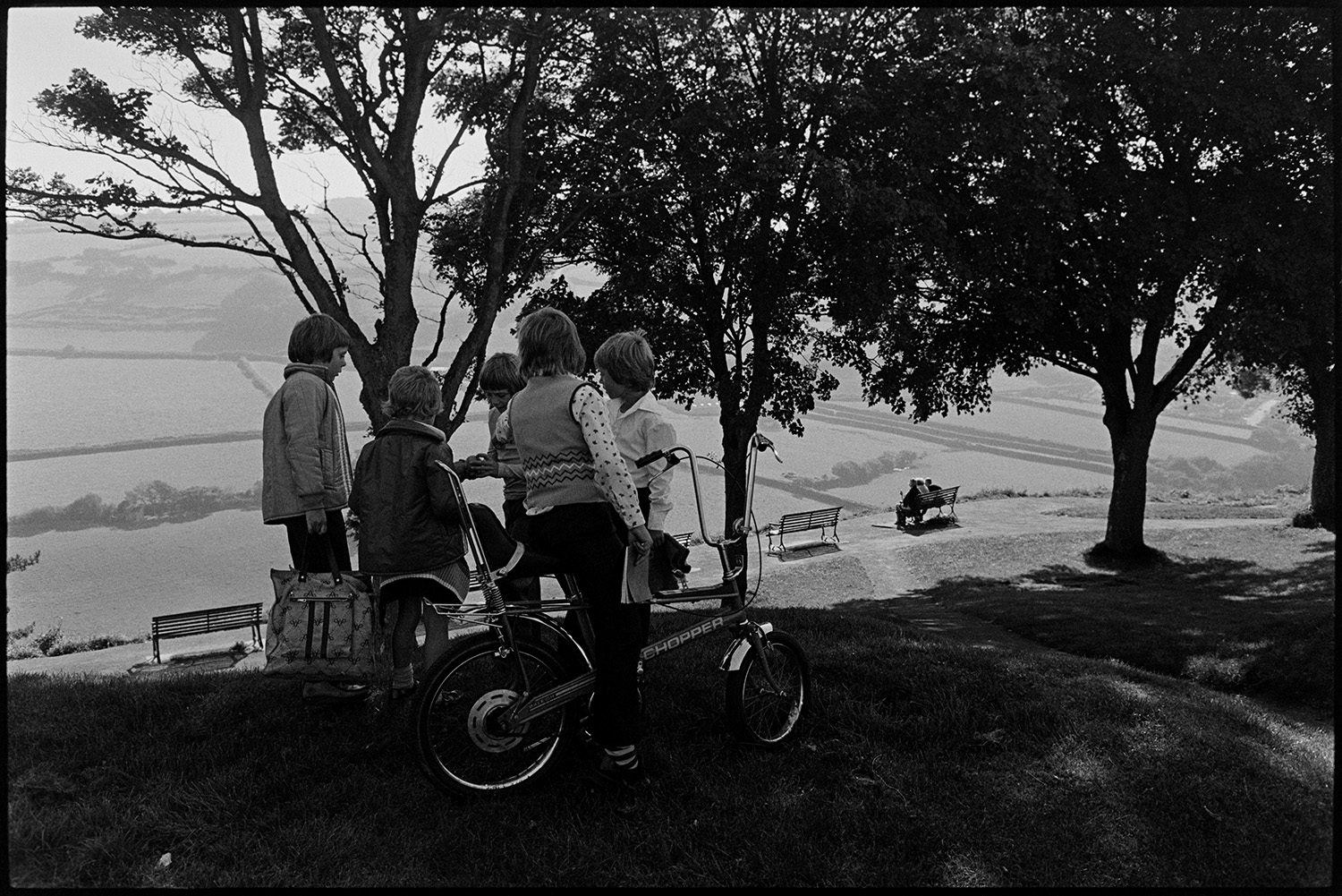 People and children enjoying park and fine view, bicycles. 
[Children playing in a park at Torrington, looking out over views of the valley below with fields and hedges. One of the boys is on a chopper bicycle. In the background people are sat on a bench looking at the view below.]