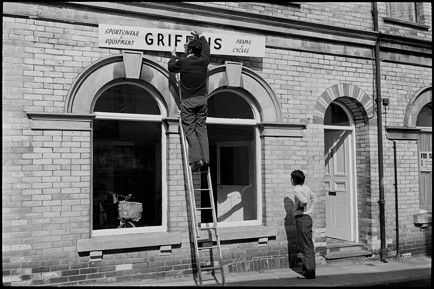 Putting up sign for new shop. 
[A man on a ladder putting up a sign for a new shop, Griffins sports shop, in Torrington. A bicycle ]