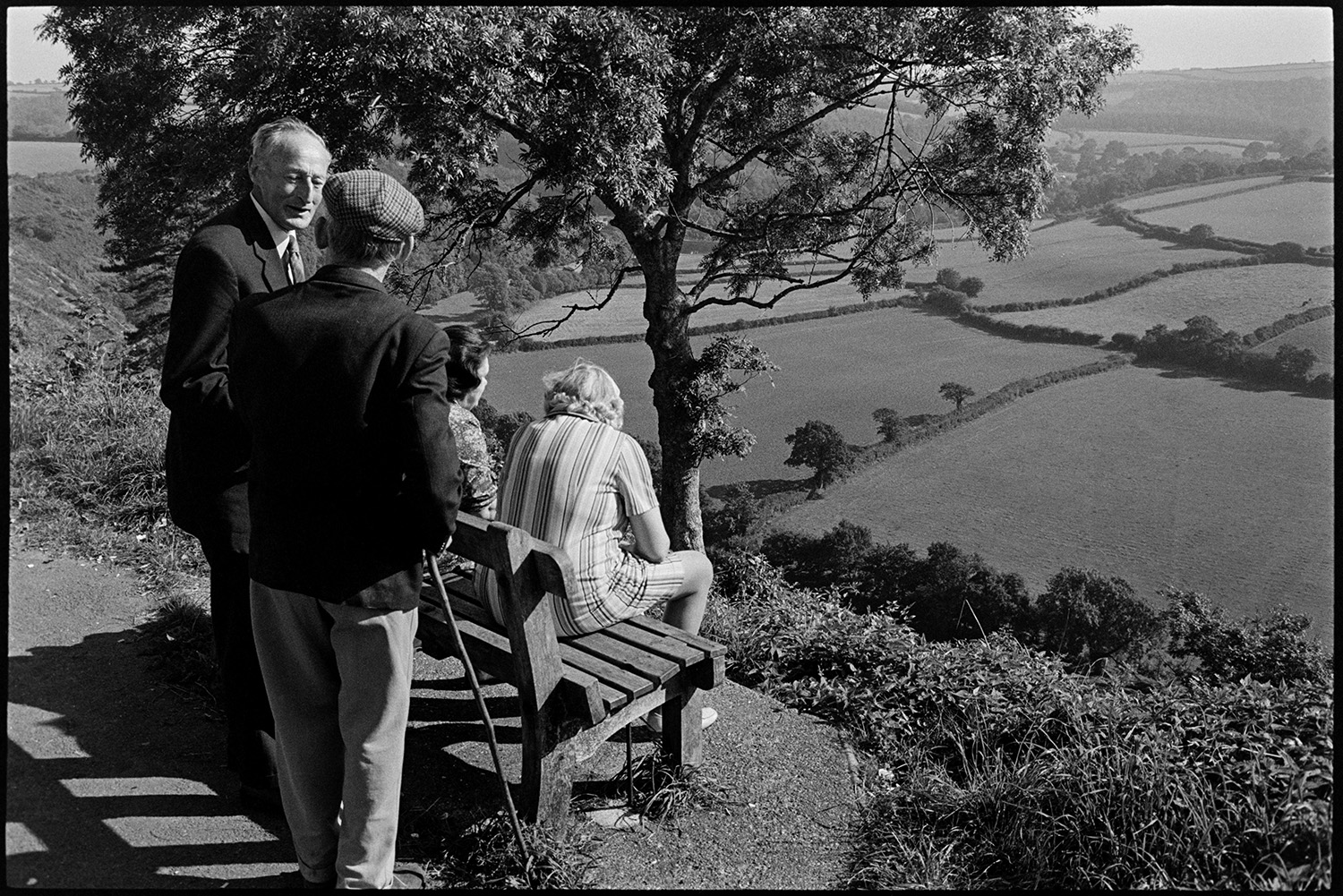 People and children enjoying park and fine view, bicycles. 
[Two women sat on a bench in a park at Torrington looking at a view of the valley below with fields, trees and hedges, Two men are stood behind the bench.]