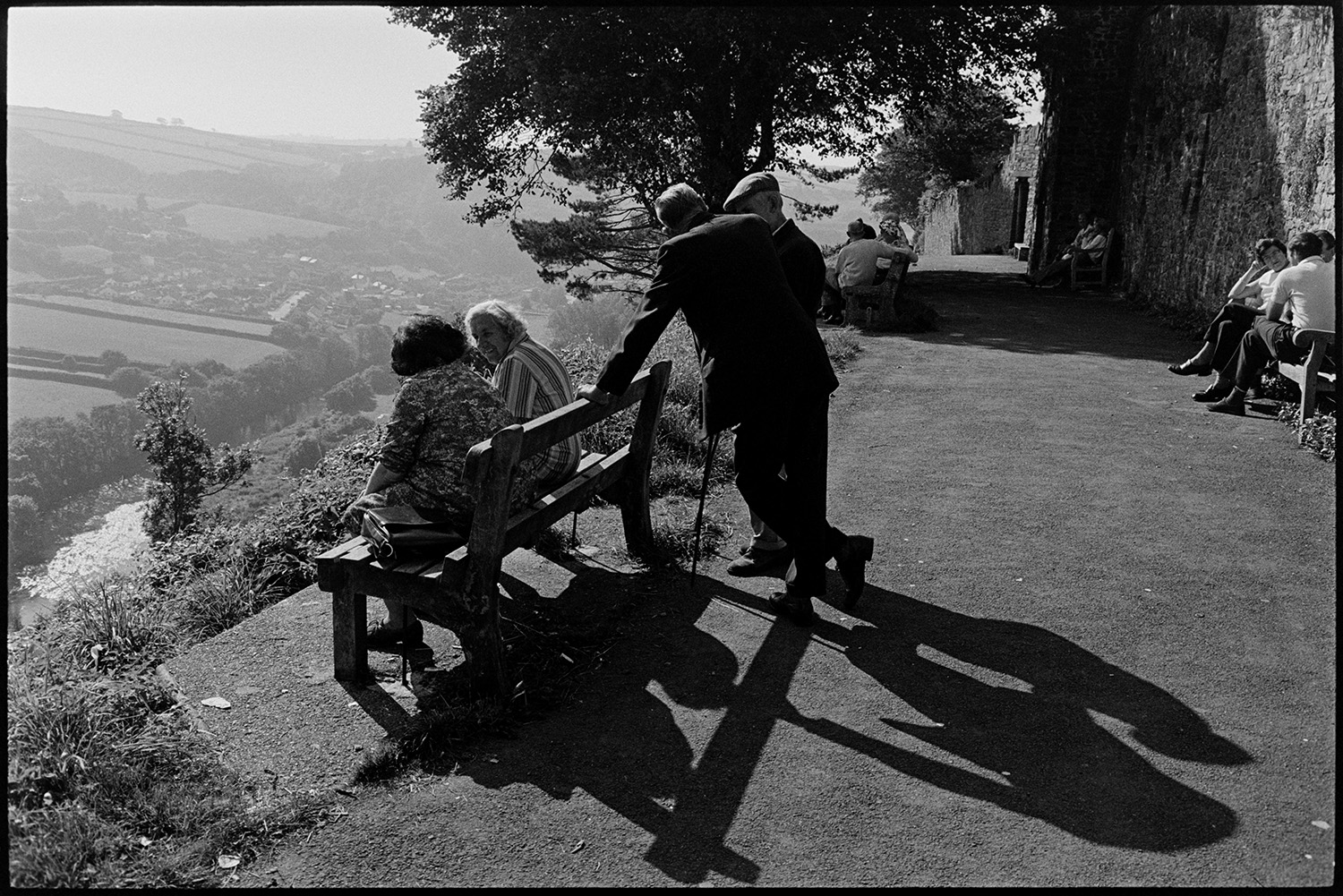 People and children enjoying park and fine view, bicycles. 
[People sat on benches along a path in a park at Torrington, looking out over a valley with fields, trees and the River Torridge. The town of Torrington can be seen in the distance.]