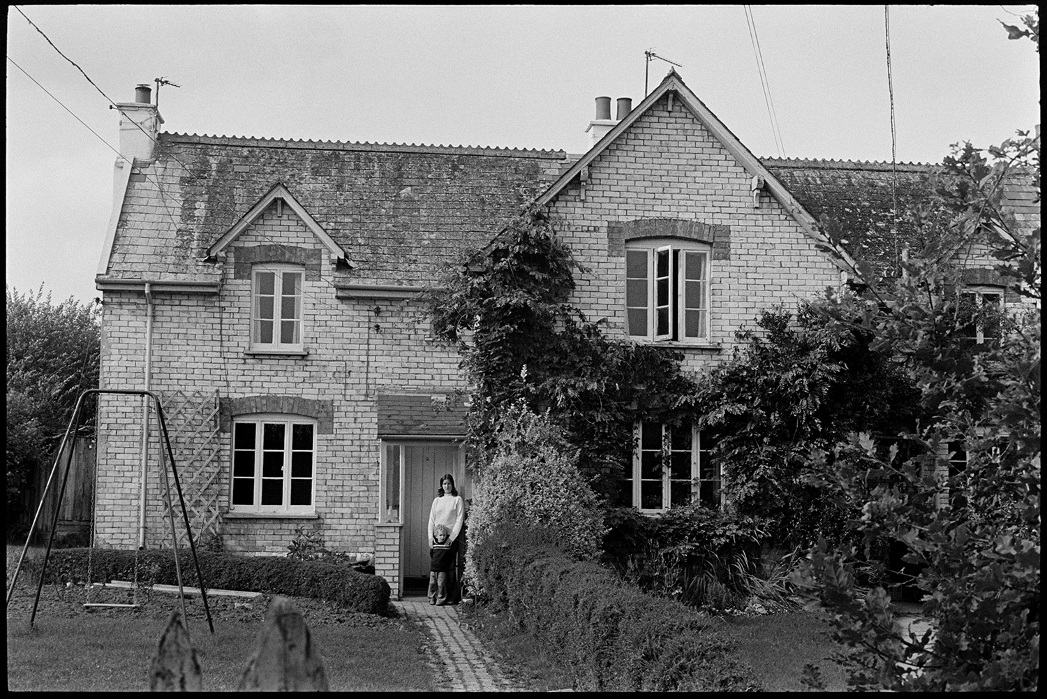 Houses taken to compare with photos in the old Archive by George Tucker. 
[A woman and a child stood outside a house in Peters Marland, Burrington. A swing can be seen in the front garden.]