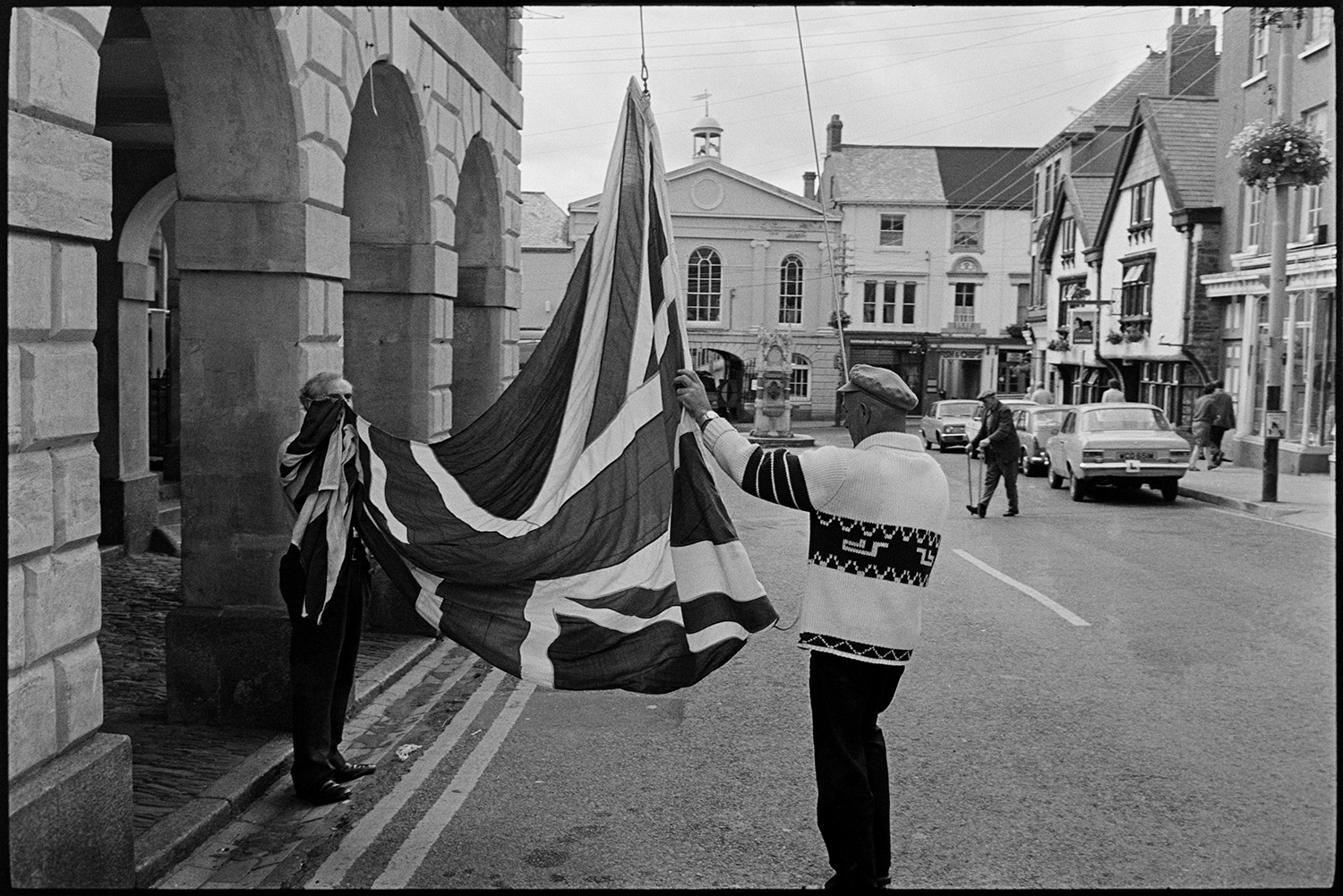 Men putting flag (Union Jack) up on Town Hall. 
[Mr Pointon and another man putting up or hoisting a Union Jack flag at the Town Hall in Torrington. Shop fronts, parked cars and shoppers can be seen in the background.]