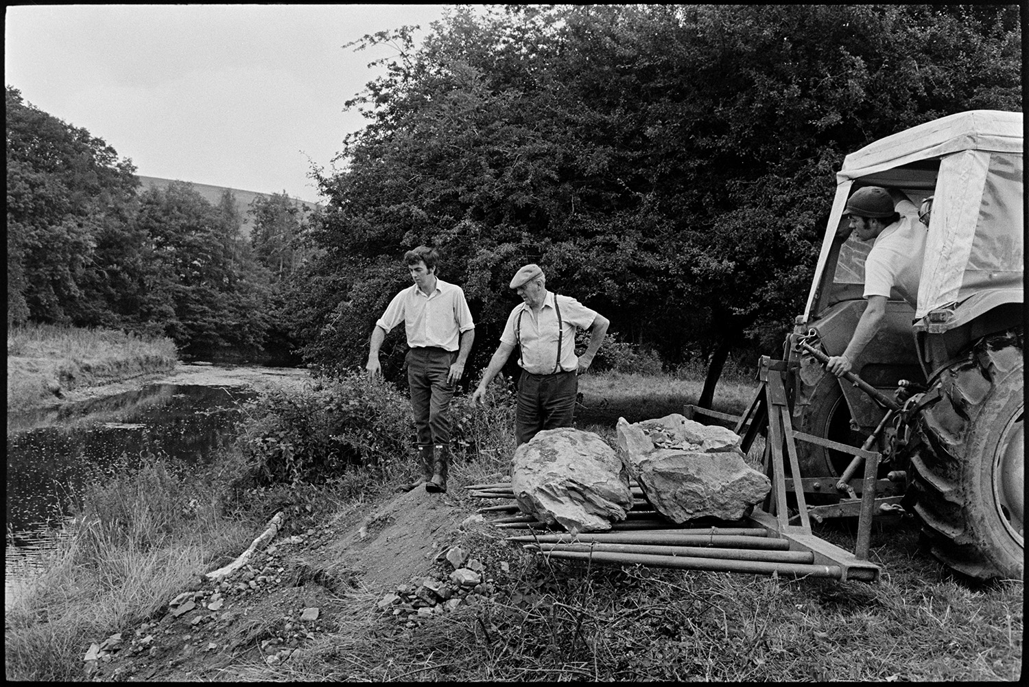 Farmers putting boulders to protect river bank. 
[John Ward and his two sons, Graham Ward and David Ward, moving boulders to a river bank using a tractor, to protect the bank from erosion, at Parsonage, Iddesleigh.]