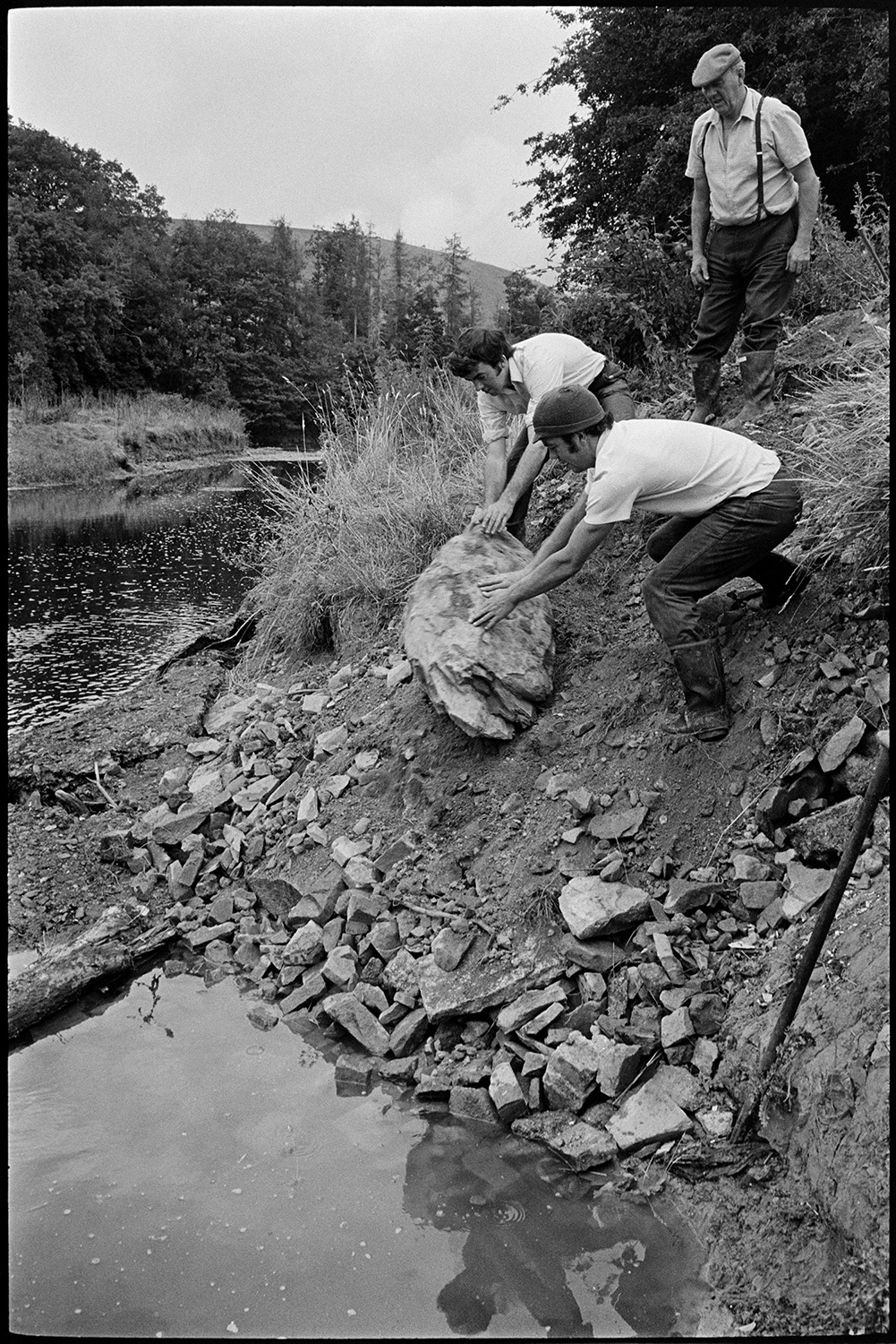 Farmers putting boulders to protect river bank. 
[John Ward and his two sons, Graham Ward and David Ward, rolling a boulder down a river bank, to protect the bank from erosion, at Parsonage, Iddesleigh.]