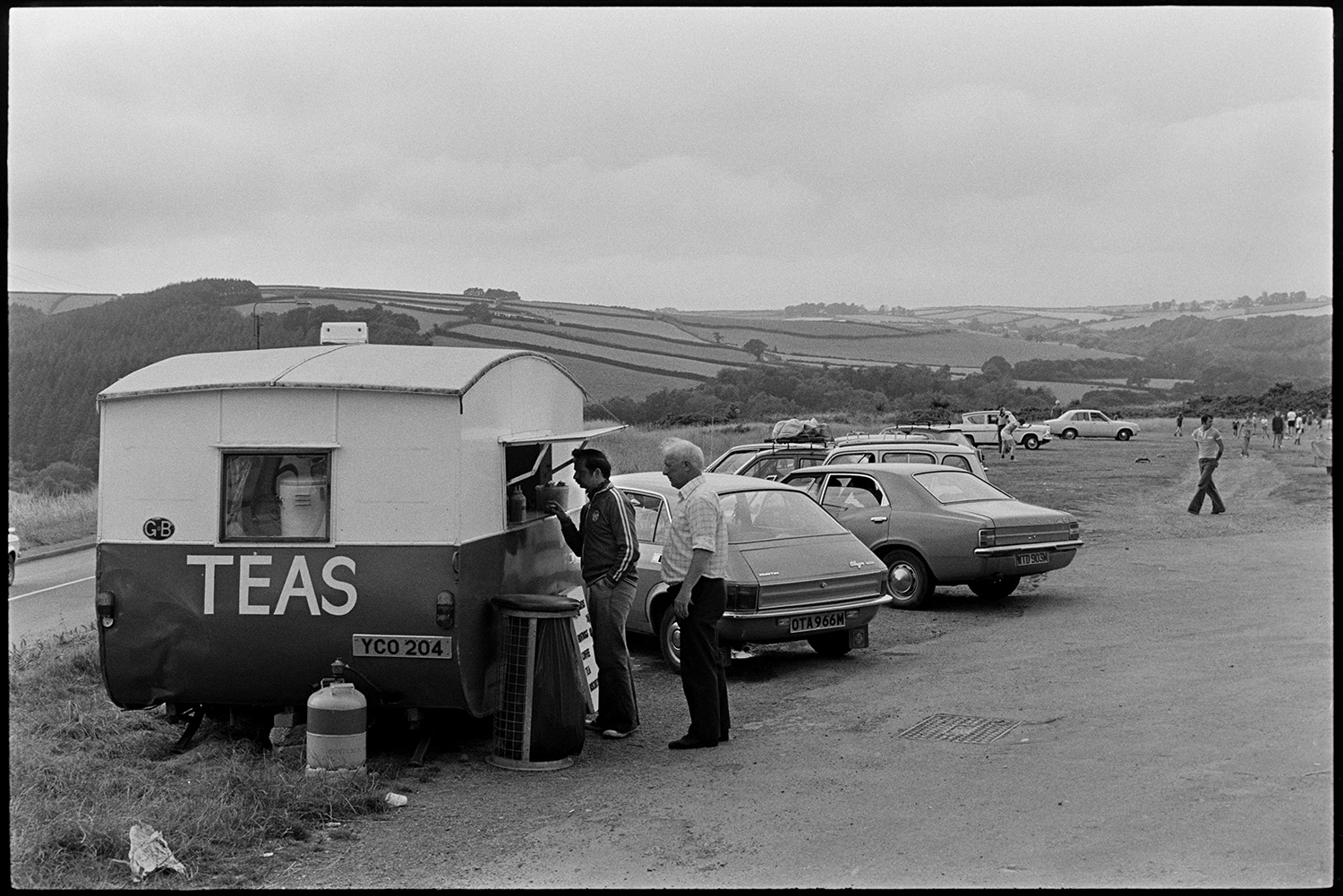 Mobile snack bar, caravan with customers in car park. 
[Two men queueing to get drinks and food from a mobile snack van , e from a converted caravan, in a car park at Torrington. Parked cars can be seen in the background and the word 'Teas' is written on the side of the snack van.]