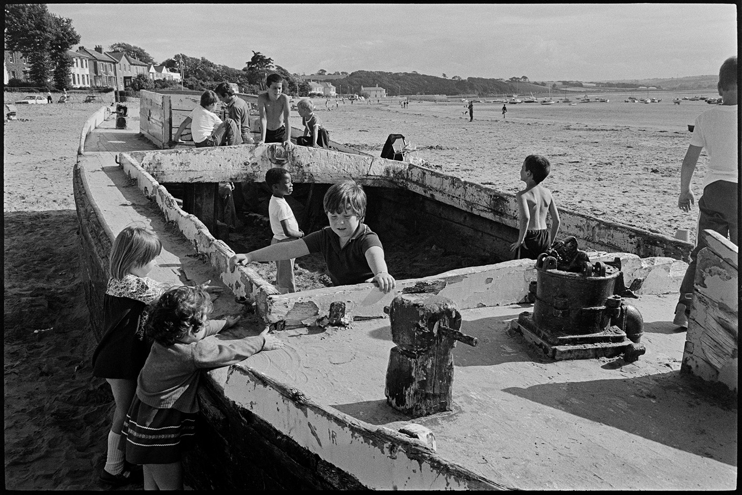 Children playing on beach, kite. Abandoned barge. 
[Children playing on an beached barge which has been abandoned, on the beach at Instow. Boats and buildings are visible in the background along the coastline.]