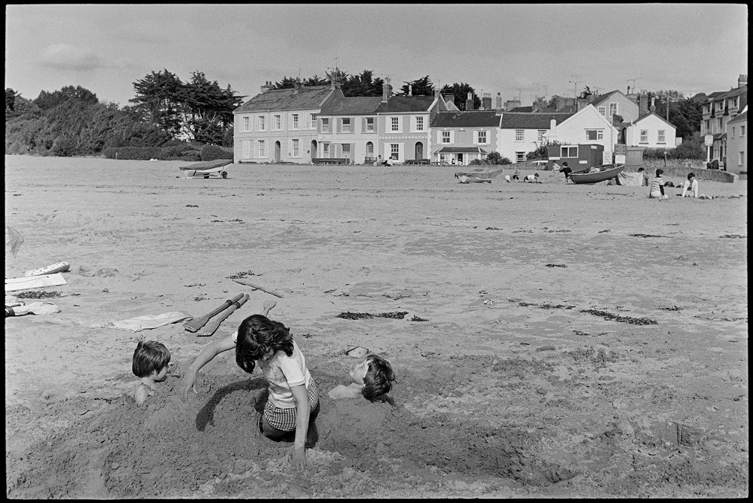 Children playing on beach, kite. Abandoned barge. 
[Children playing on the beach at Instow. They are burying themselves in the sand. Buildings and boats on the seafront are visible in the background.]