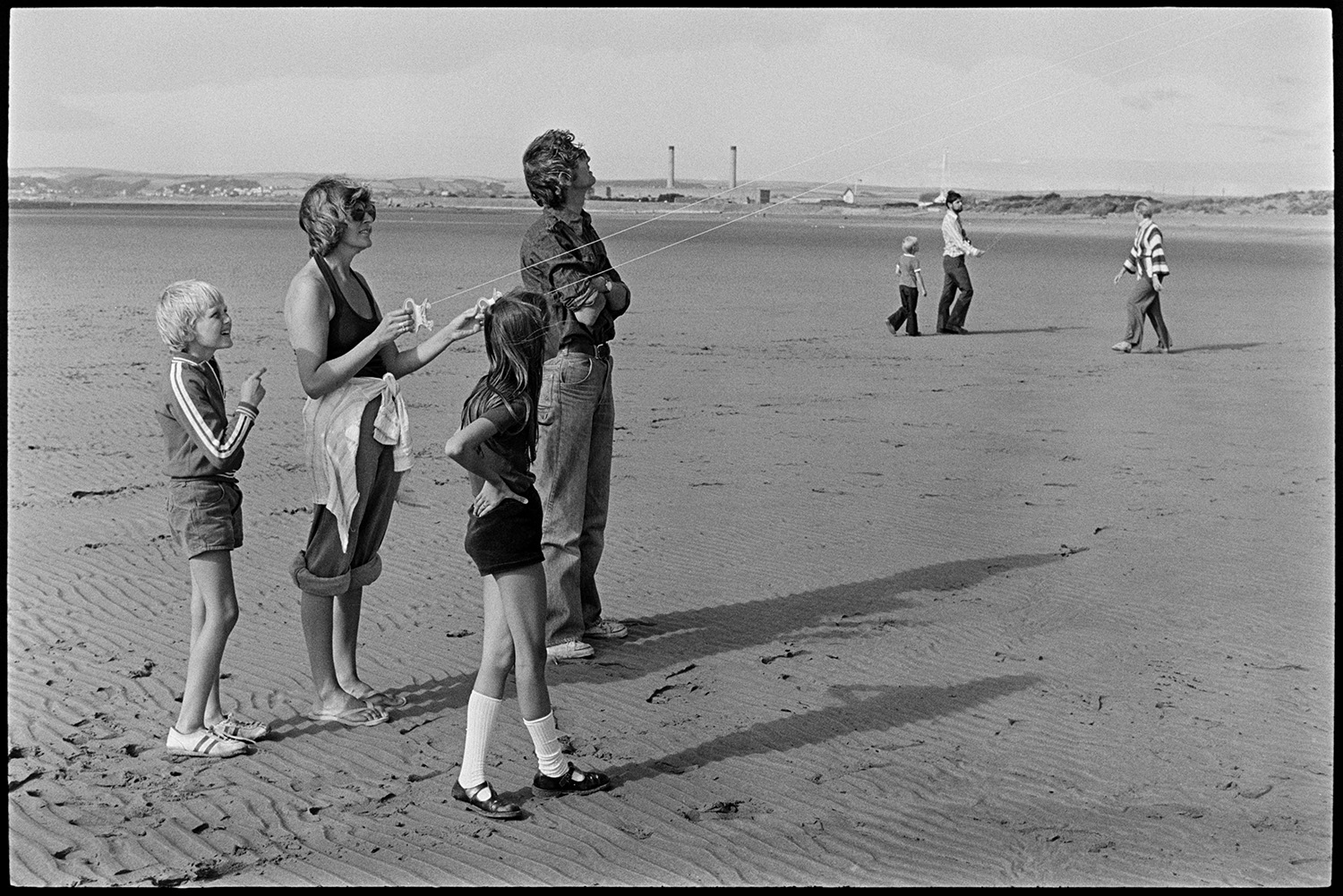 Children playing on beach, kite. Abandoned barge. 
[A family flying a kite on the beach at Instow. Sand dunes and Yelland power station are visible in the background.]