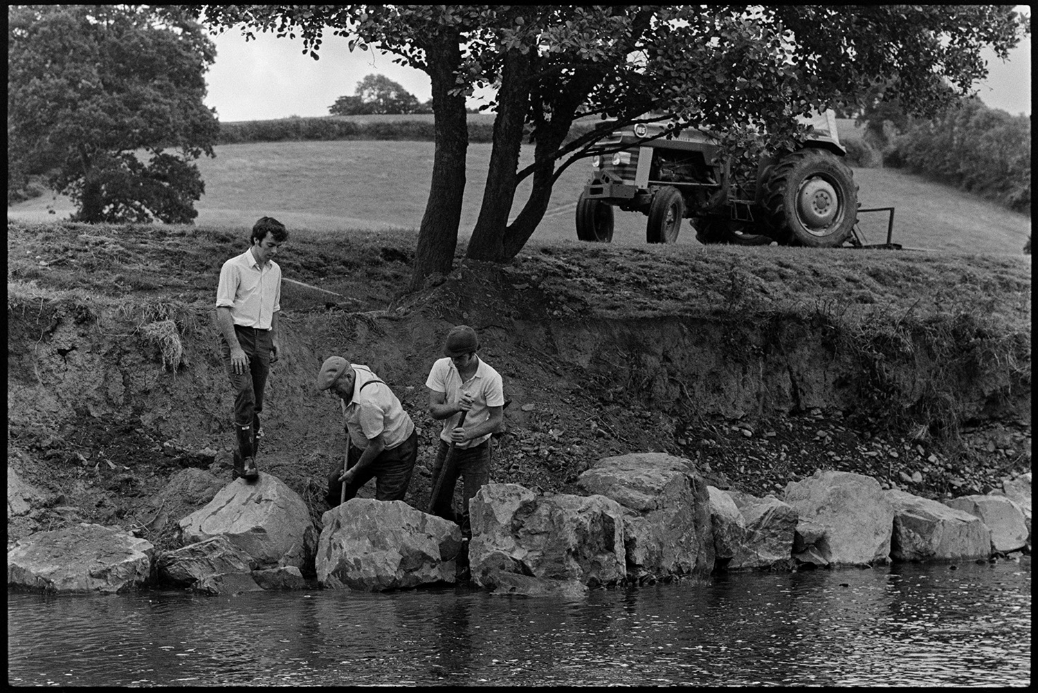 Farmers putting boulders to protect river bank. 
[John Ward and his two sons, Graham Ward and David Ward, moving boulders with crow bars by a river bank, to protect the bank from erosion , at Parsonage, Iddesleigh. The tractor they have used to transport the boulders can be seen in a field in the background.]