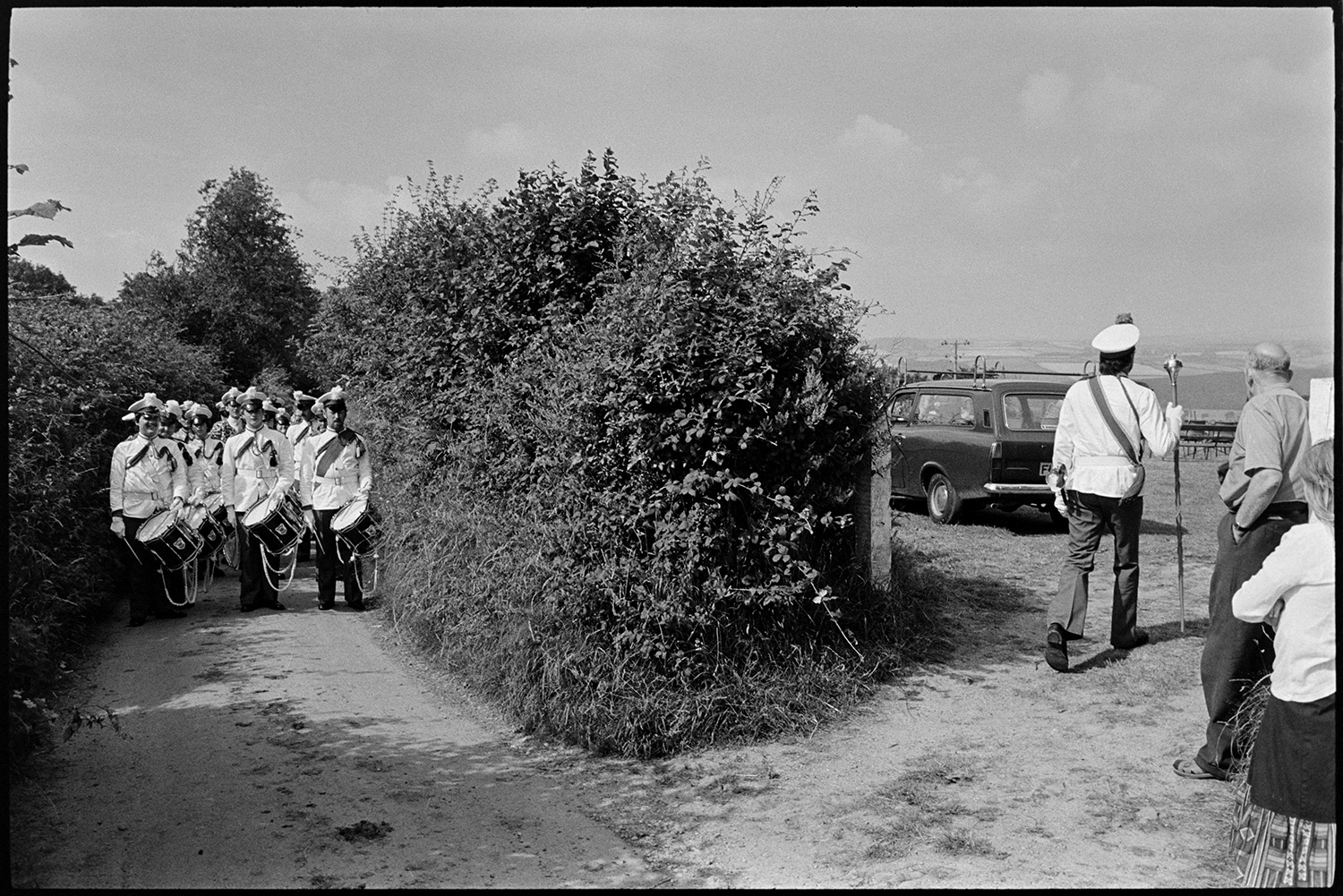 Band and drum majorettes on parade. 
[A band leader holding a mace and a drum band marching along a lane and into a field for Atherington Village fete.]