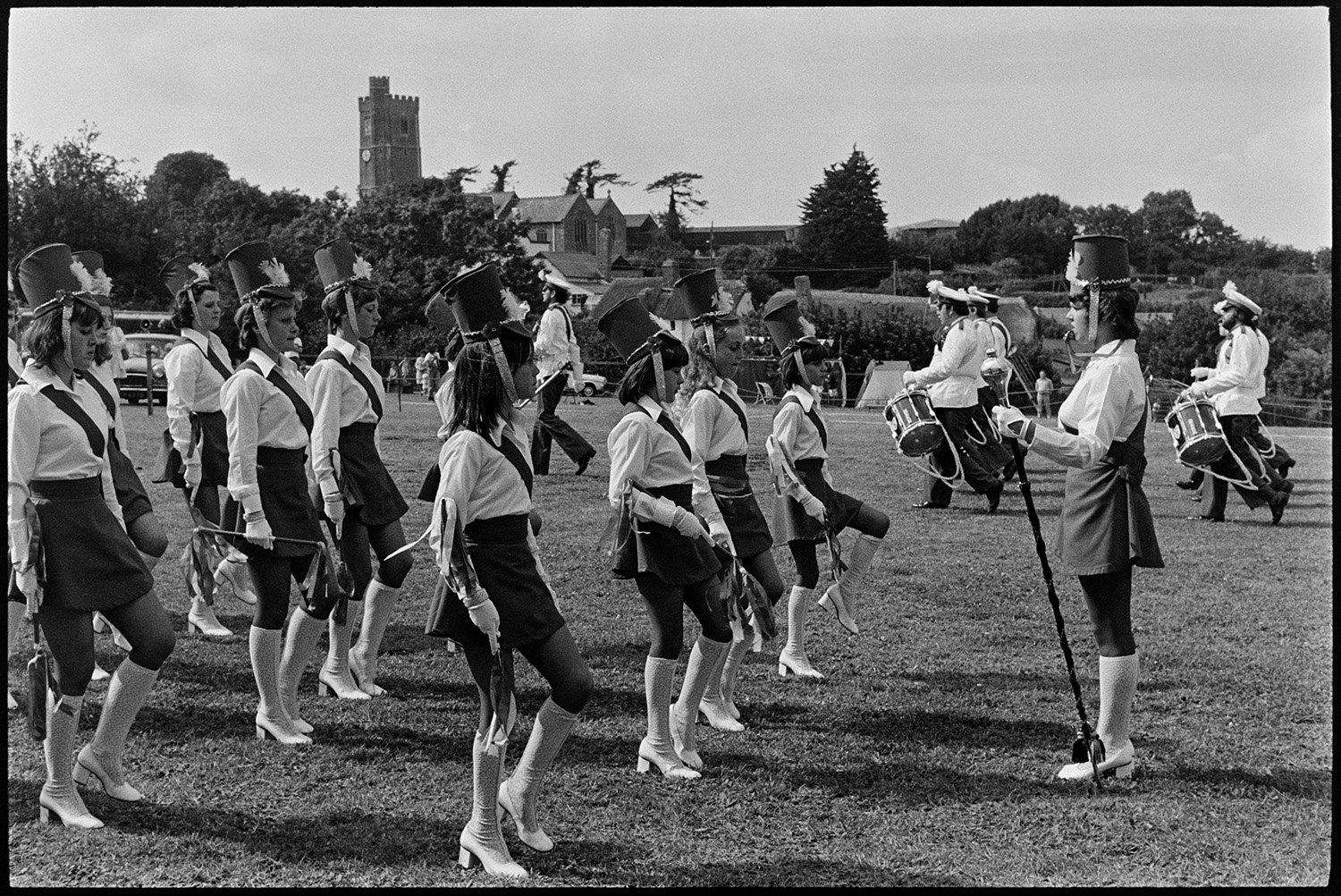 Band and drum majorettes on parade. 
[Majorettes performing at Atherington Village fete. A drum band are marching past in the background.]