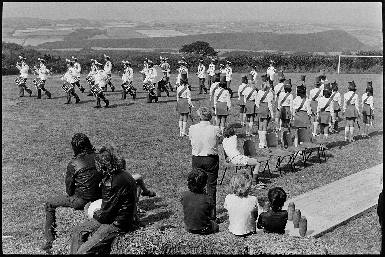 Band and drum majorettes on parade. 
[Majorettes and a drum band performing at Atherington Village fete. People are watching from a skittle alley set up by hay bales.]