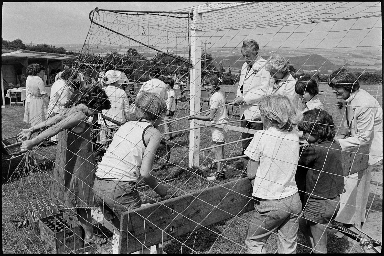 Amusements at village fete flowers, pony rides, skittles etc. 
[Men, women and children playing a fishing game in a goal net at Atherington Village fete. Boxes of drinks bottles can be seen on the ground by the game. Other people are looking at stalls in the background.]